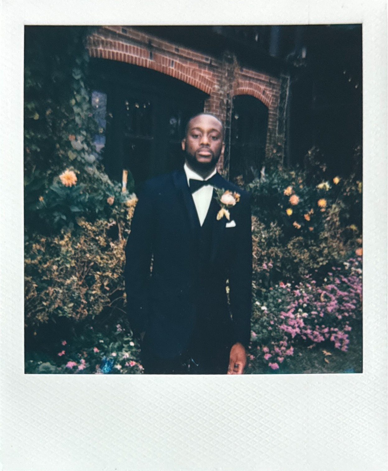 Man in a black tuxedo with a bow tie and boutonnière, standing in front of a brick building with flowers and greenery. Gramercy Mansion Maryland Wedding Film Photographer.