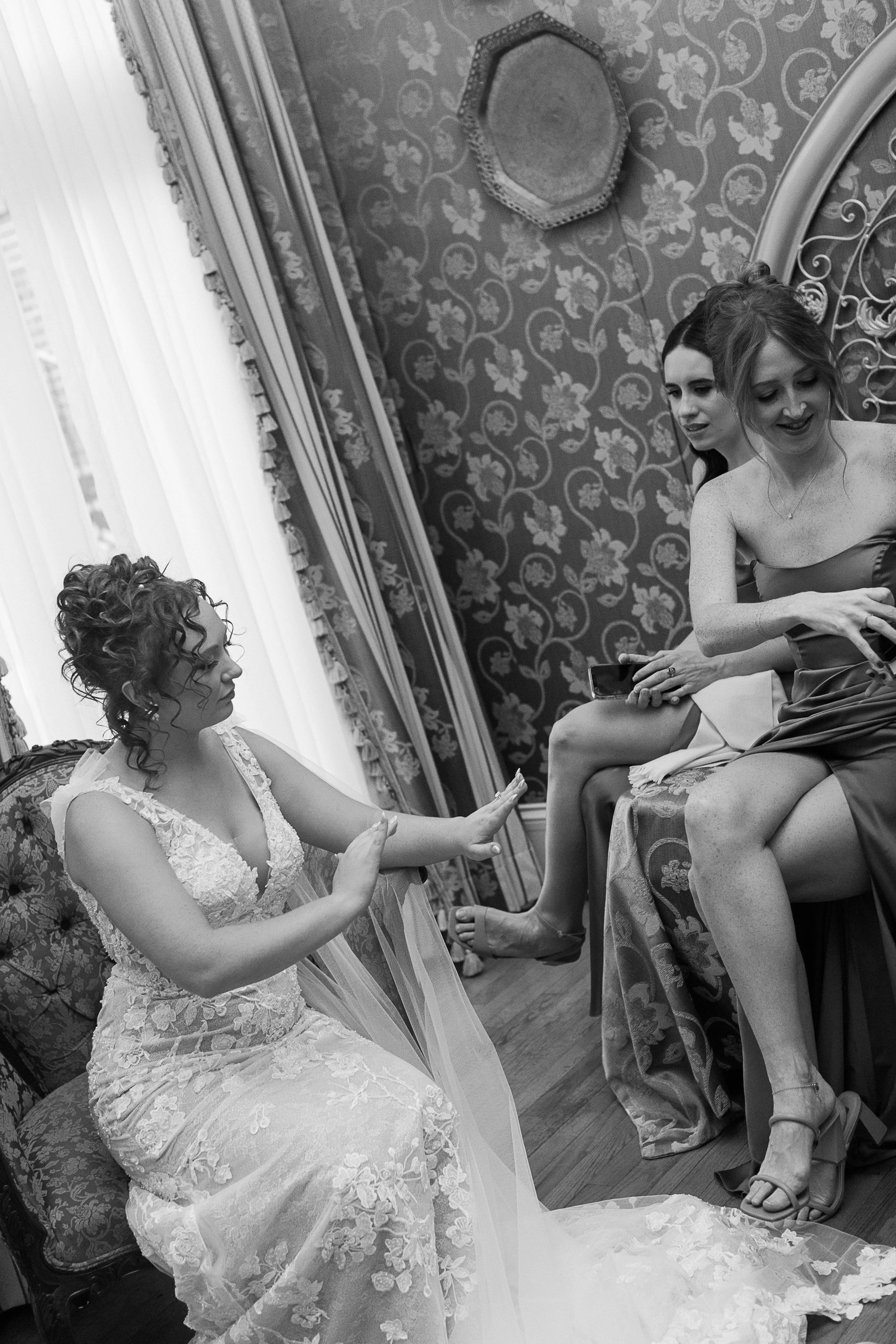 A bride in a lace wedding dress sits on a chair while adjusting her veil, with two women seated next to her on a decorated seat. The room features patterned wallpaper and curtains. gramercy mansion maryland wedding