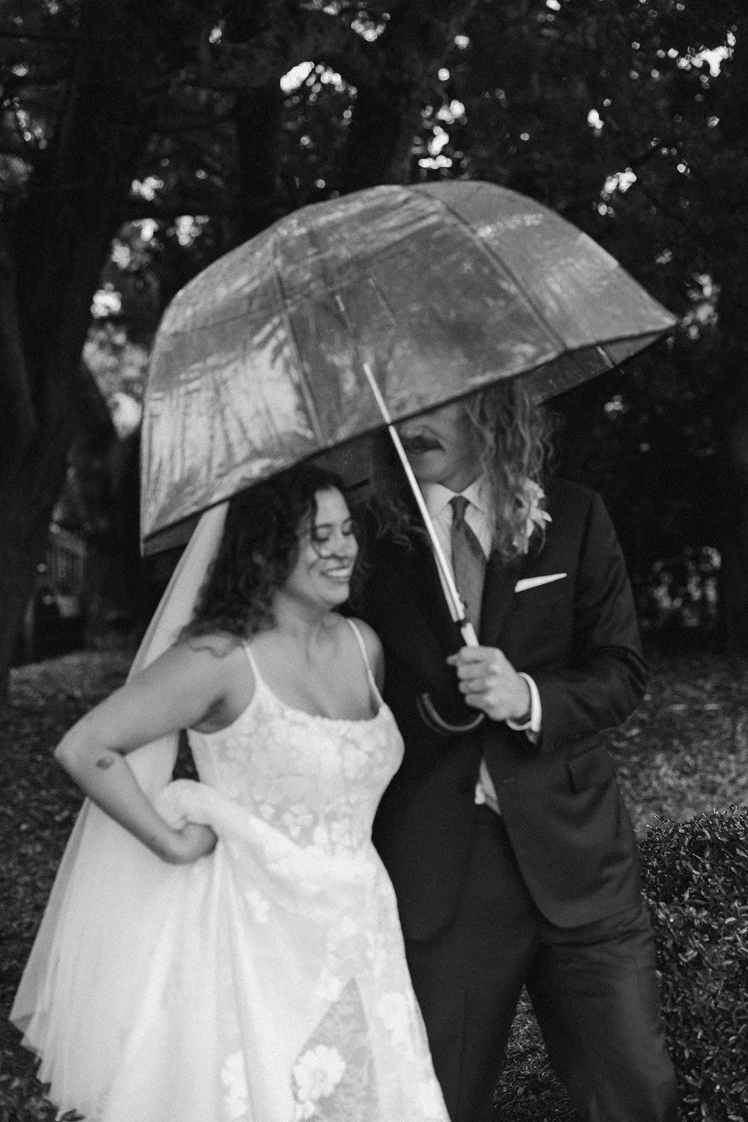 A bride and groom standing close together under a large umbrella, smiling and looking at each other, outdoors in a park setting. ocracoke outerbanks wedding film photographer