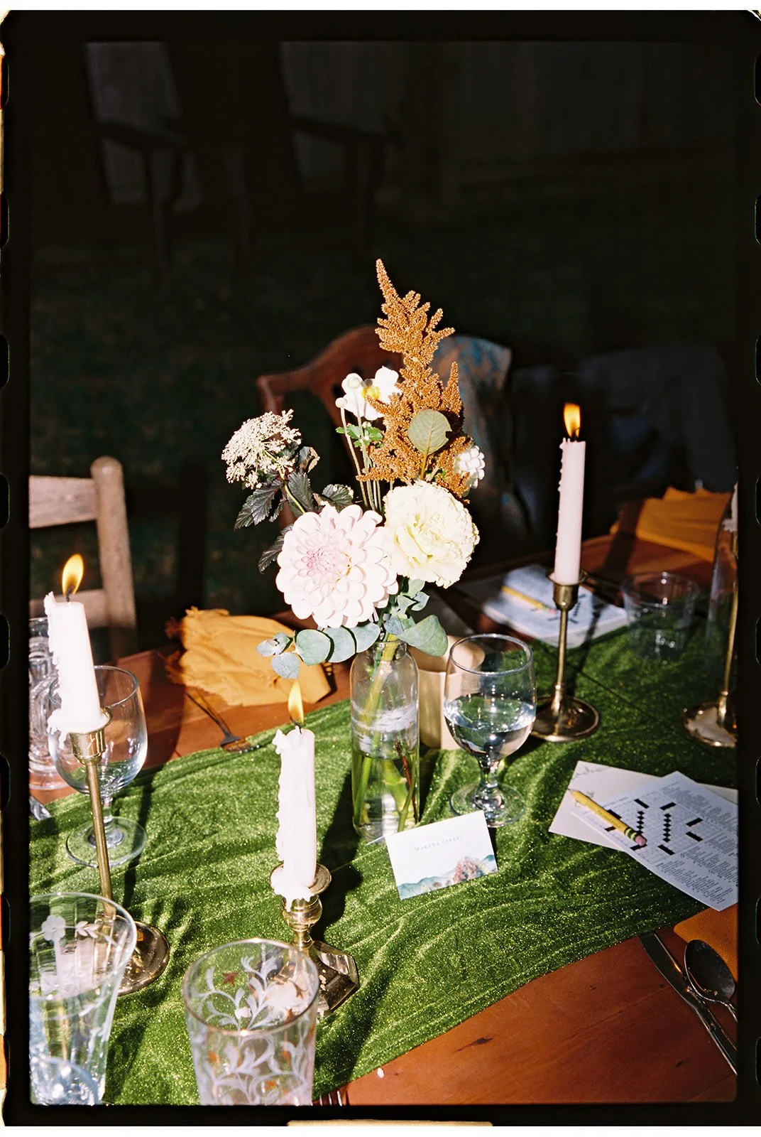 A decorated dinner table with a green table runner, brass candlesticks with white candles, a clear glass vase with white and pink flowers, and glasses and menus. The table is set for a formal event or celebration. Roanoke Virginia Wedding Film Photog