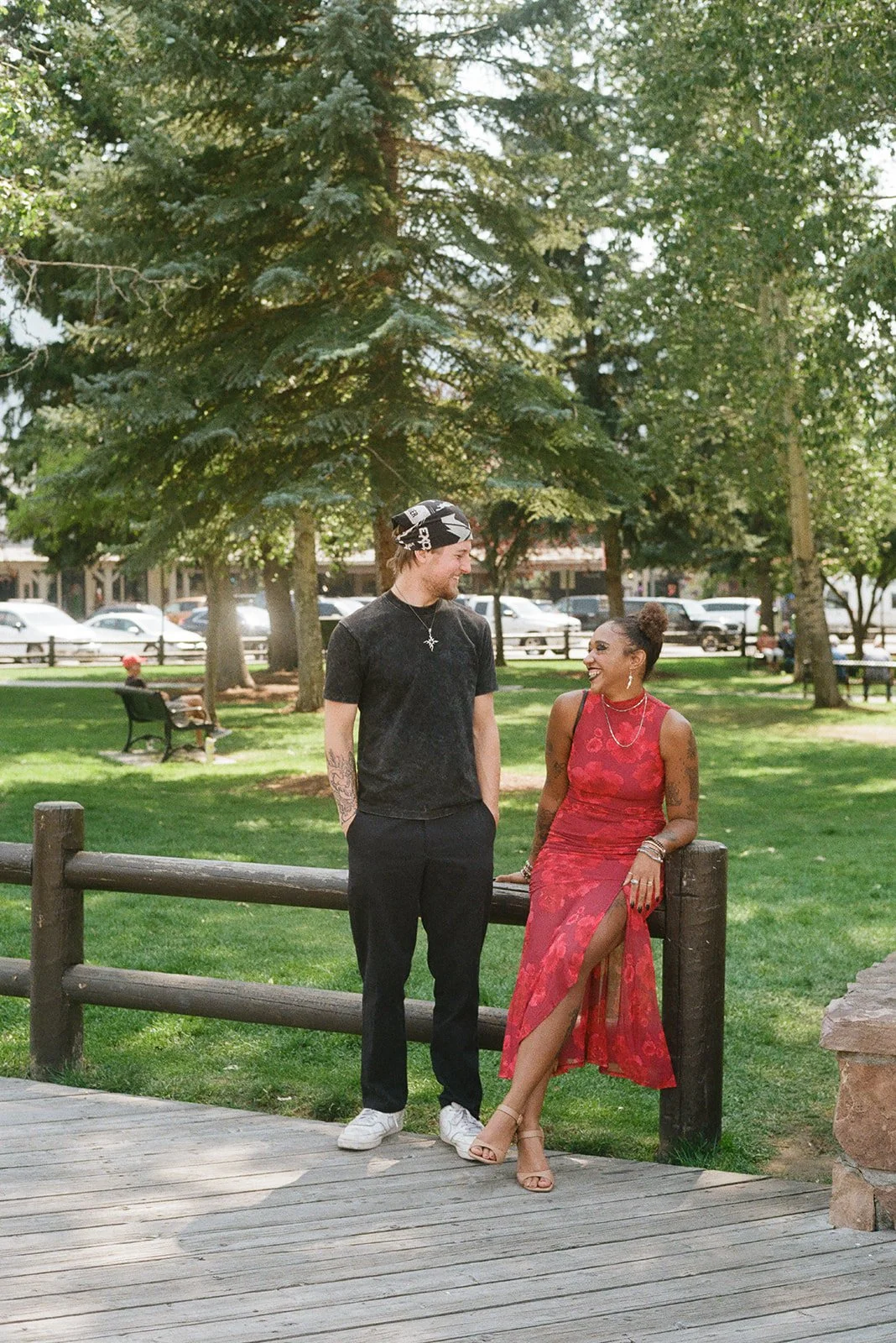 A young man and woman are standing and sitting on a wooden railing in a park, engaging in conversation, surrounded by trees and greenery, with cars and benches visible in the background. Jackson Wyoming Engagement Shoot
