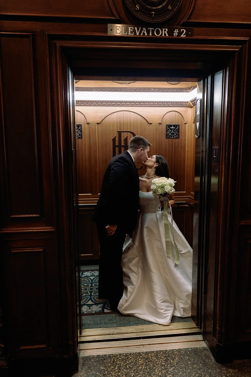 A bride and groom sharing a kiss inside a vintage wooden elevator, with the bride holding a bouquet of white roses, and a sign above the elevator door reading 'ELEVATOR #2'. roanoke wedding film photographer
