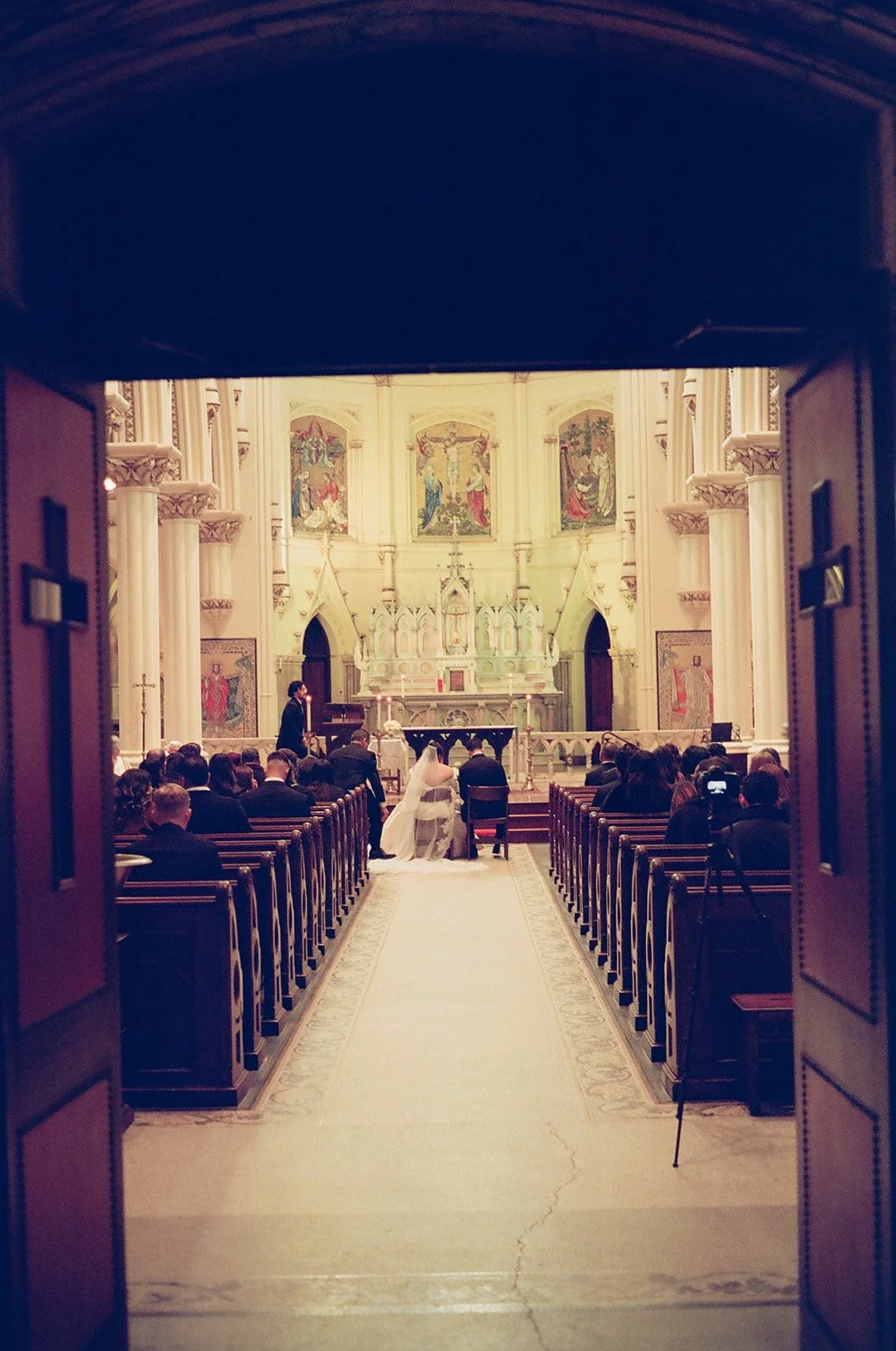 View through church doors of wedding ceremony inside a church with guests, bride, groom, and organist, ornate altar, and religious artworks. Baltimore Maryland Wedding Film Photographer