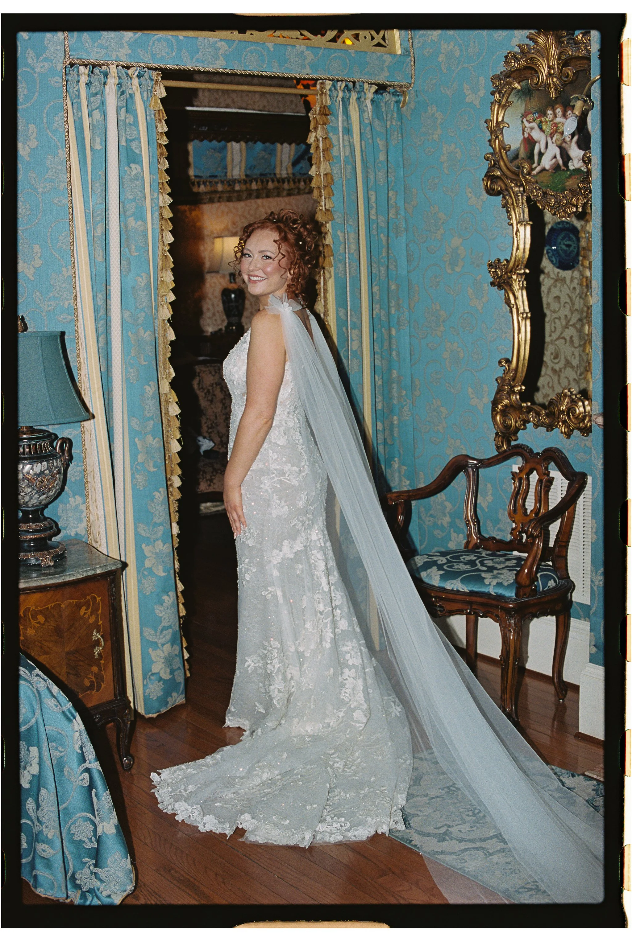 Bride standing in an ornate room wearing a lace wedding gown with a long veil, surrounded by blue patterned drapery and vintage furniture. gramercy mansion maryland wedding