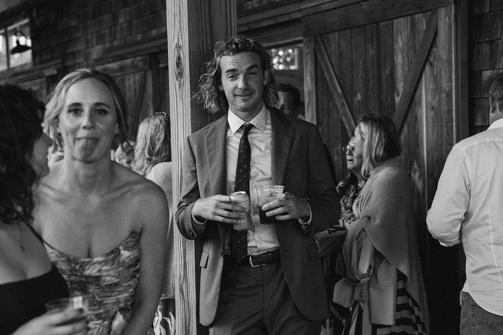 People socializing at a gathering, with a man in a suit holding drinks in the center, a woman sticking out her tongue on the left, and others engaged in conversation at a rustic venue. ocracoke outerbanks wedding film photographer