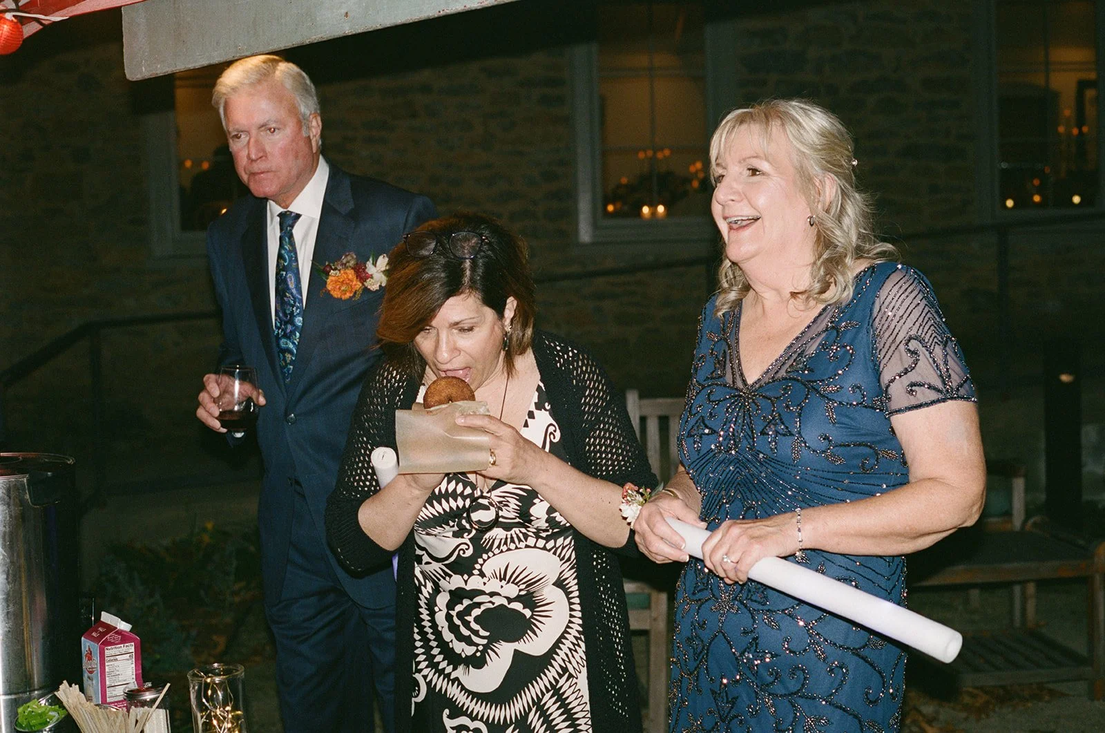 Three adults in formal attire at a social event, with one woman eating a food item from a paper container, holding a rolled-up paper, and the other two standing nearby. The background shows a rustic brick wall with windows. potters craft cider