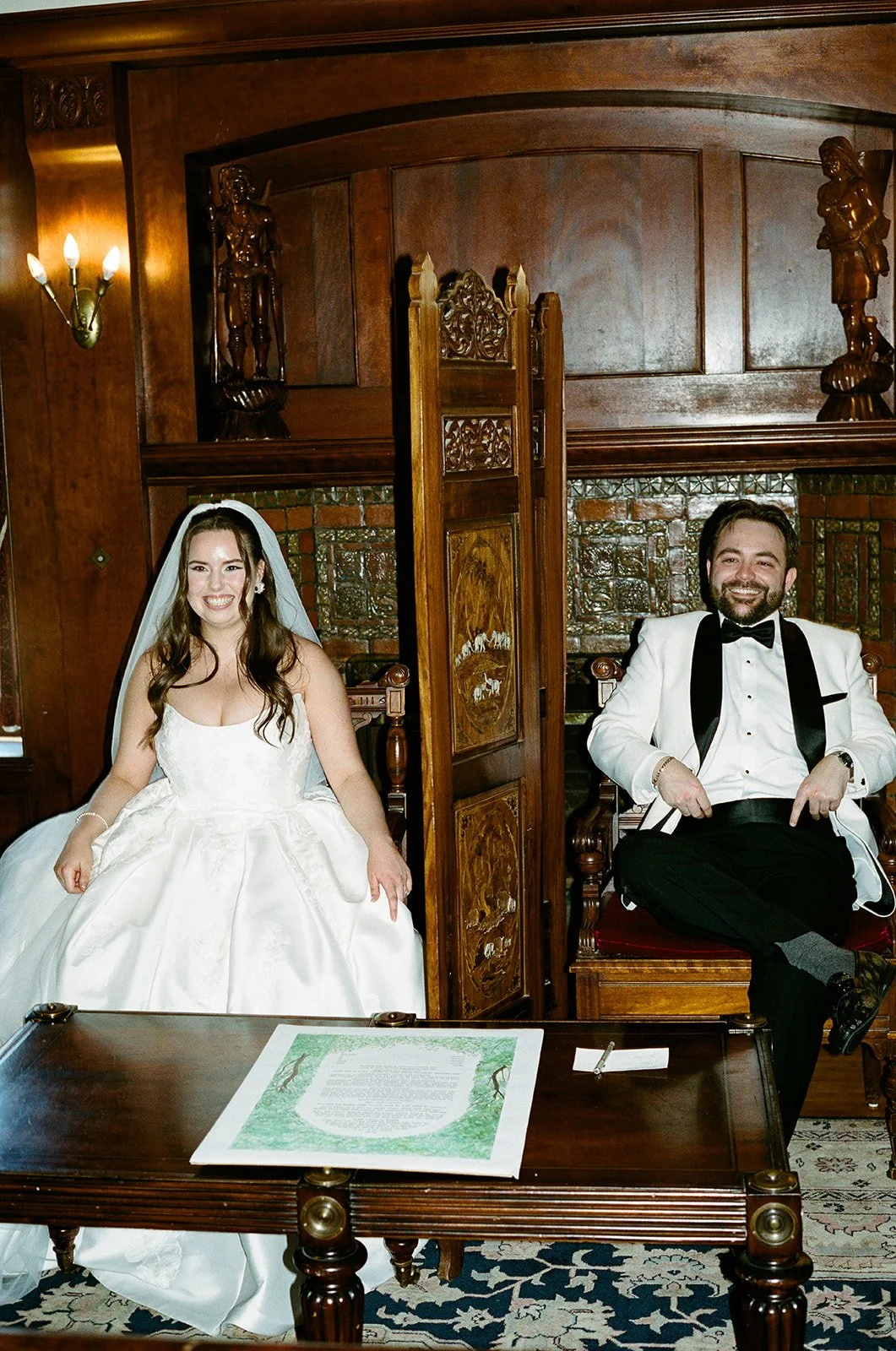 A bride and groom seated inside a wood-paneled room, smiling during their wedding ceremony. The bride is in a white wedding gown and veil, and the groom is in a white tuxedo jacket with a black bow tie. Gramercy Mansion Baltimore Maryland Photograph