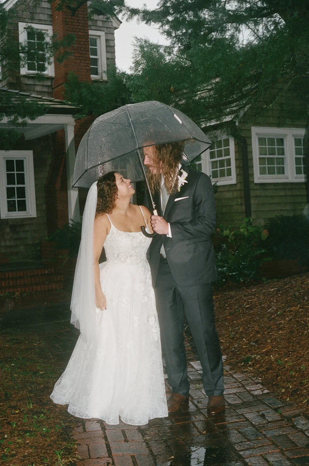 A bride and groom stand close under a large umbrella on a rain-soaked brick walkway outside a house, gazing into each other's eyes. ocracoke outerbanks wedding film photographer