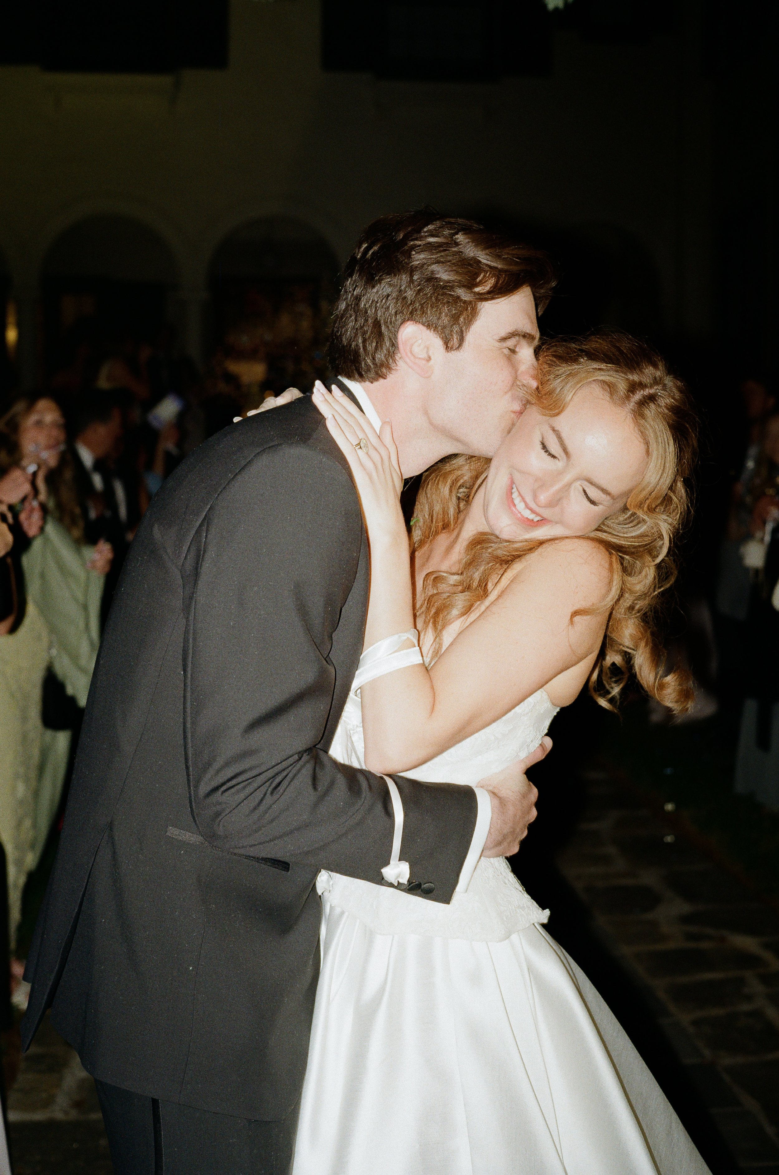 A groom kissing a bride on her forehead during a wedding reception, with guests in the background.
