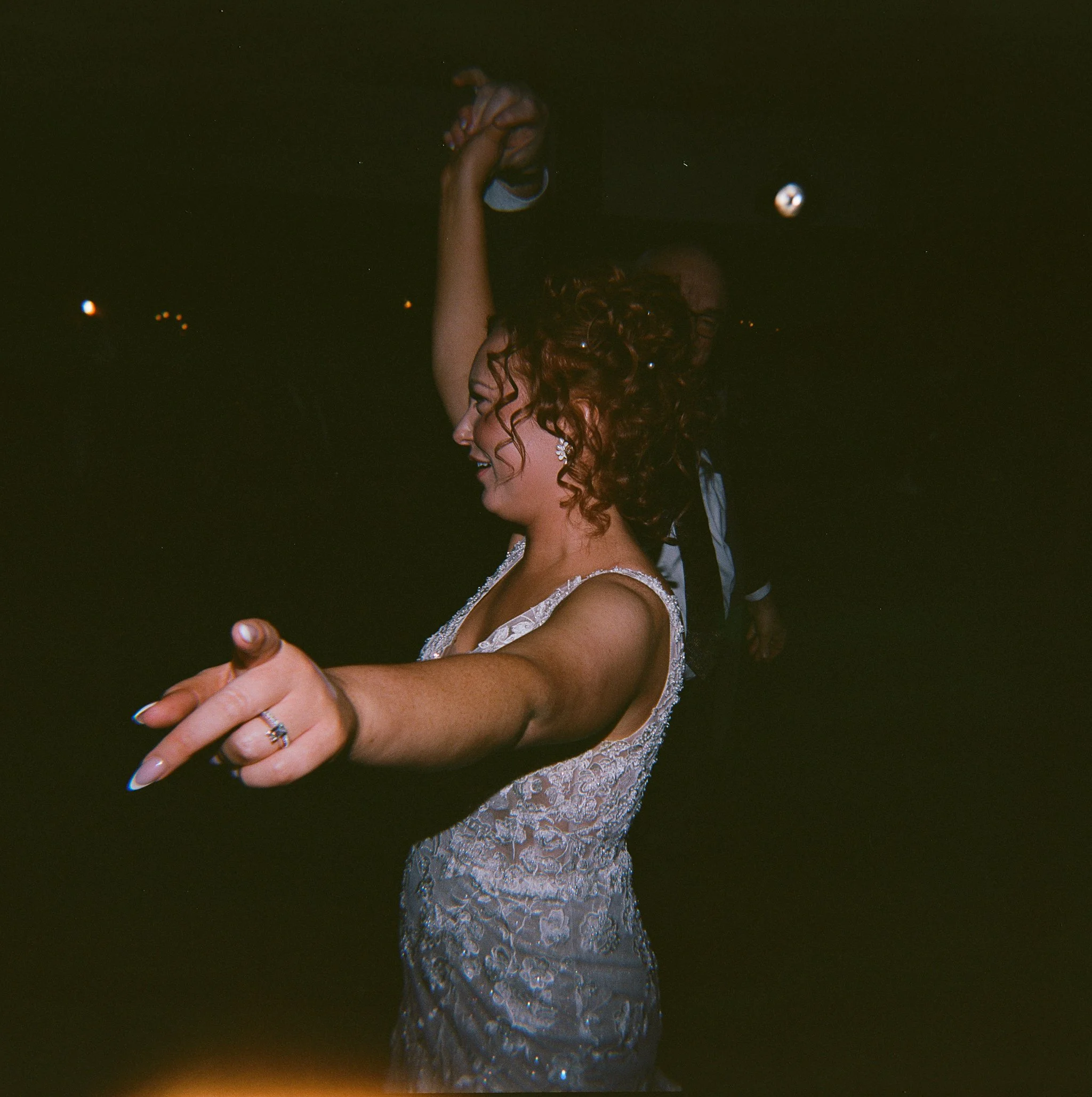 A bride dancing with a partner in a dark room, wearing a lace wedding gown and earrings, smiling with an outstretched arm. gramercy mansion maryland wedding holga 120 film