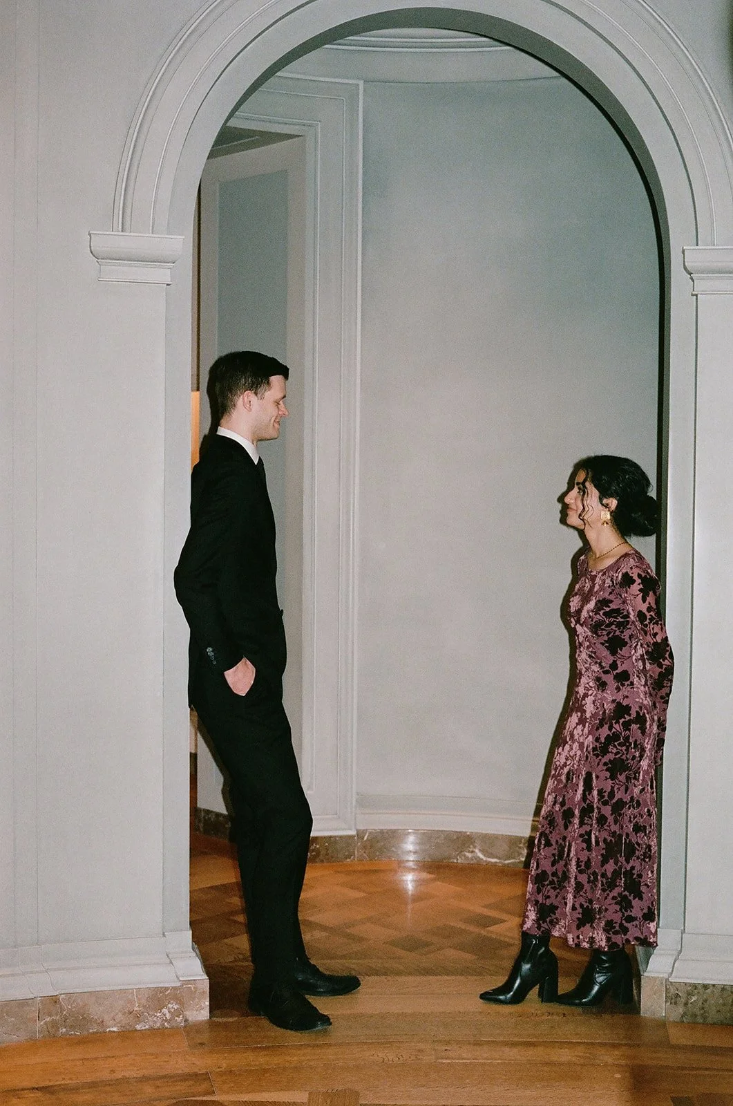 A man and a woman standing in front of a large archway, facing each other and smiling, in an indoor setting with wood flooring and light-colored walls. washington dc gallery of art Wedding Film Photographer