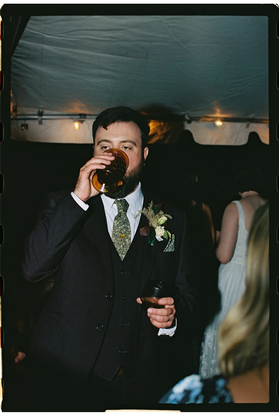 A man in a suit with a boutonniere drinks from a glass at a social event, with other people in the background. Roanoke Virginia Wedding Film Photographer.