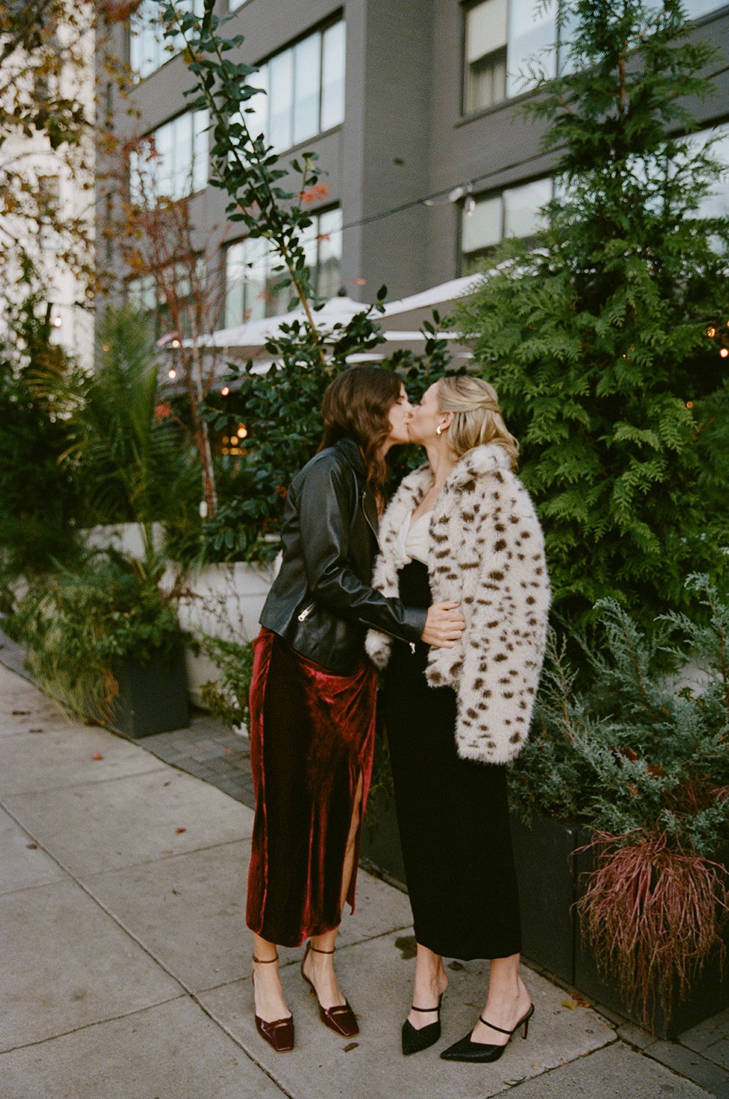 Two women kiss on a city sidewalk during the evening, surrounded by greenery and buildings in the background. DC Wedding film photographer doubles cafe