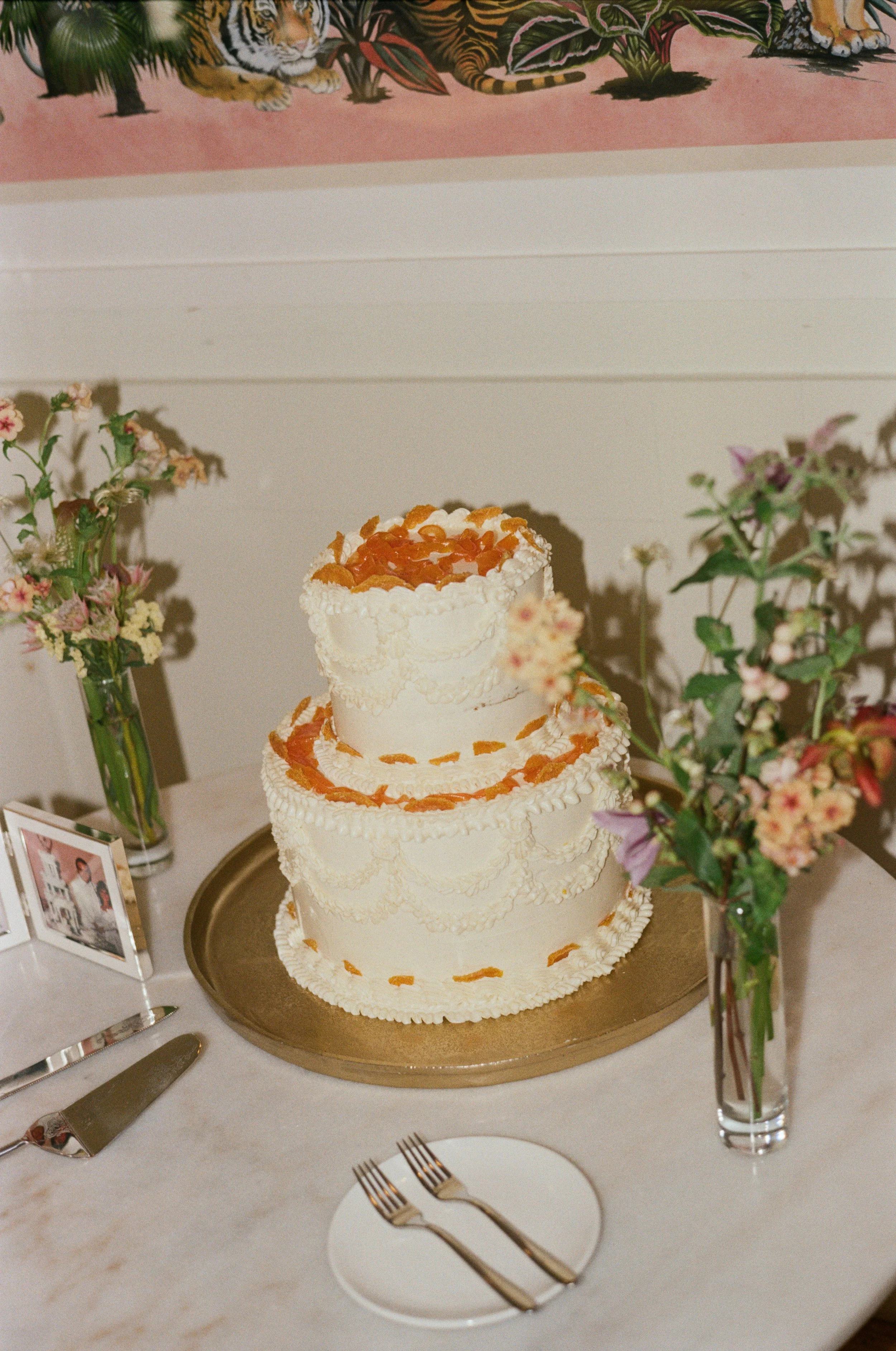 Three-tiered white wedding cake decorated with orange and white frosting details, on a gold tray, surrounded by pink and white flowers in vases, with a small photo frame and silver cake knife and forks on a marble table at Common House in Richmond VA