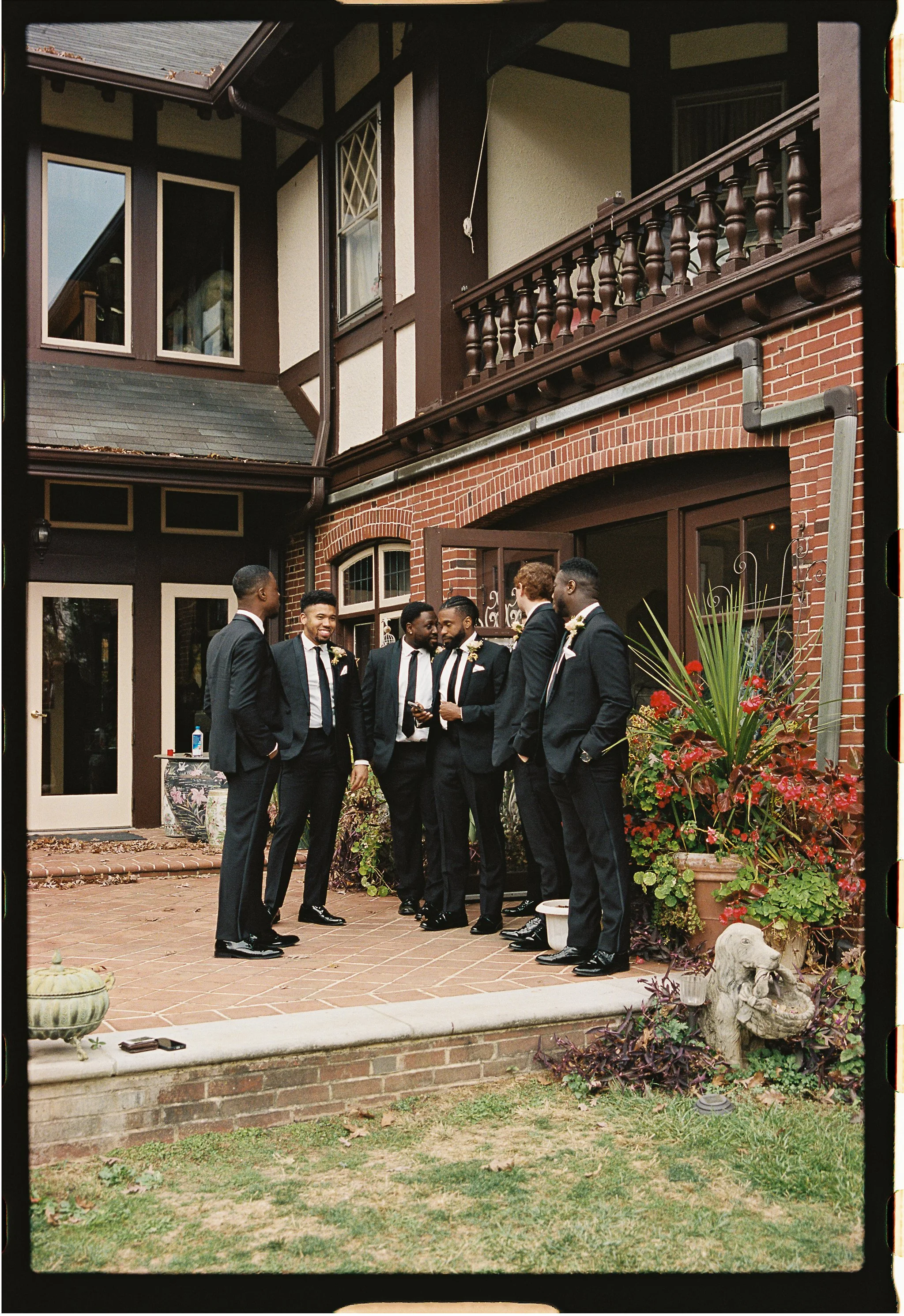 Group of groomsmen wearing suits with boutonnieres, standing and talking on a brick patio outside a Tudor-style house. There are decorative plants and a small sculpture nearby. gramercy mansion maryland wedding