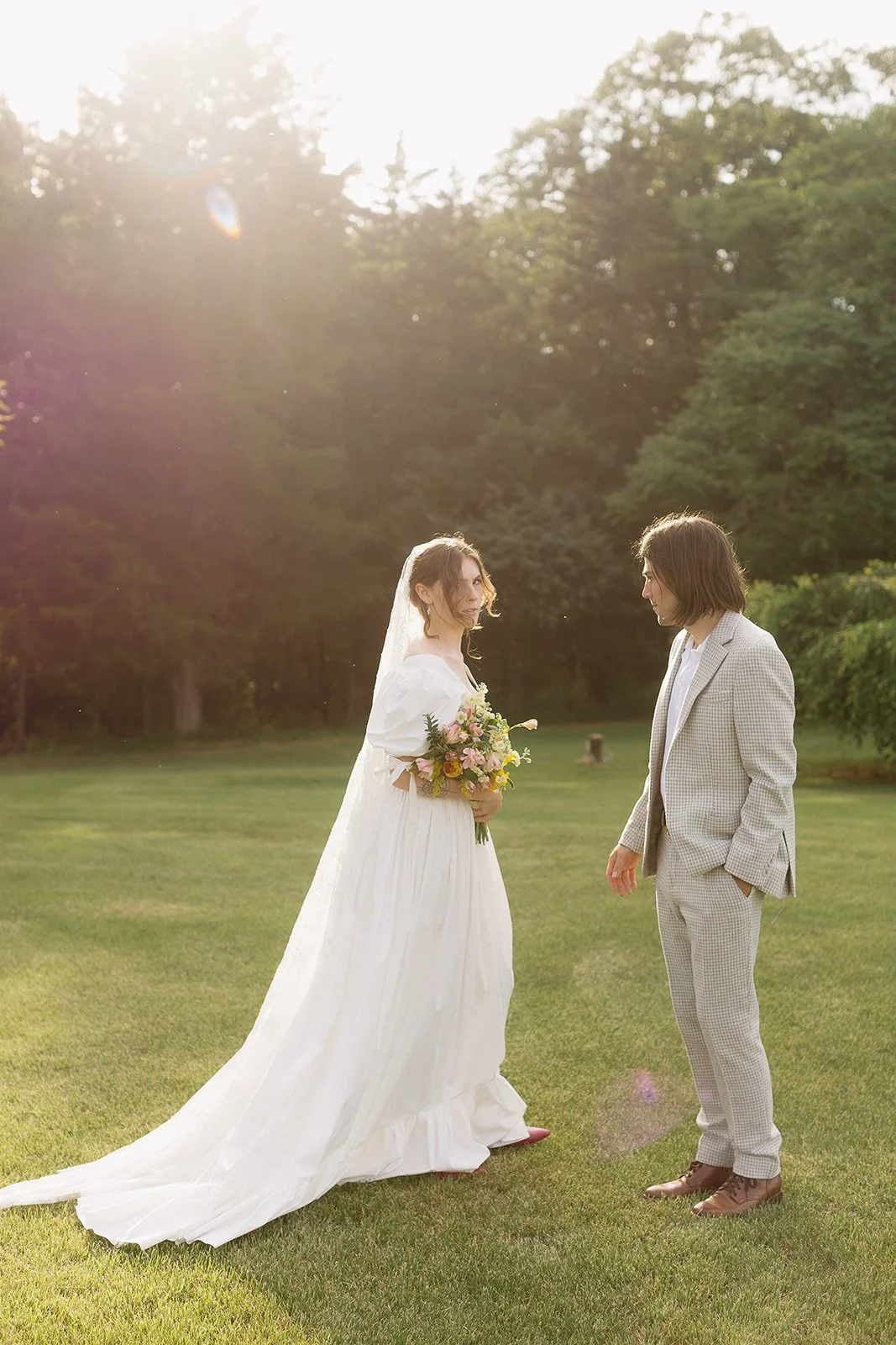 Bride and groom standing on grass in sunlight; bride wearing white dress and holding bouquet, groom in light plaid suit.
