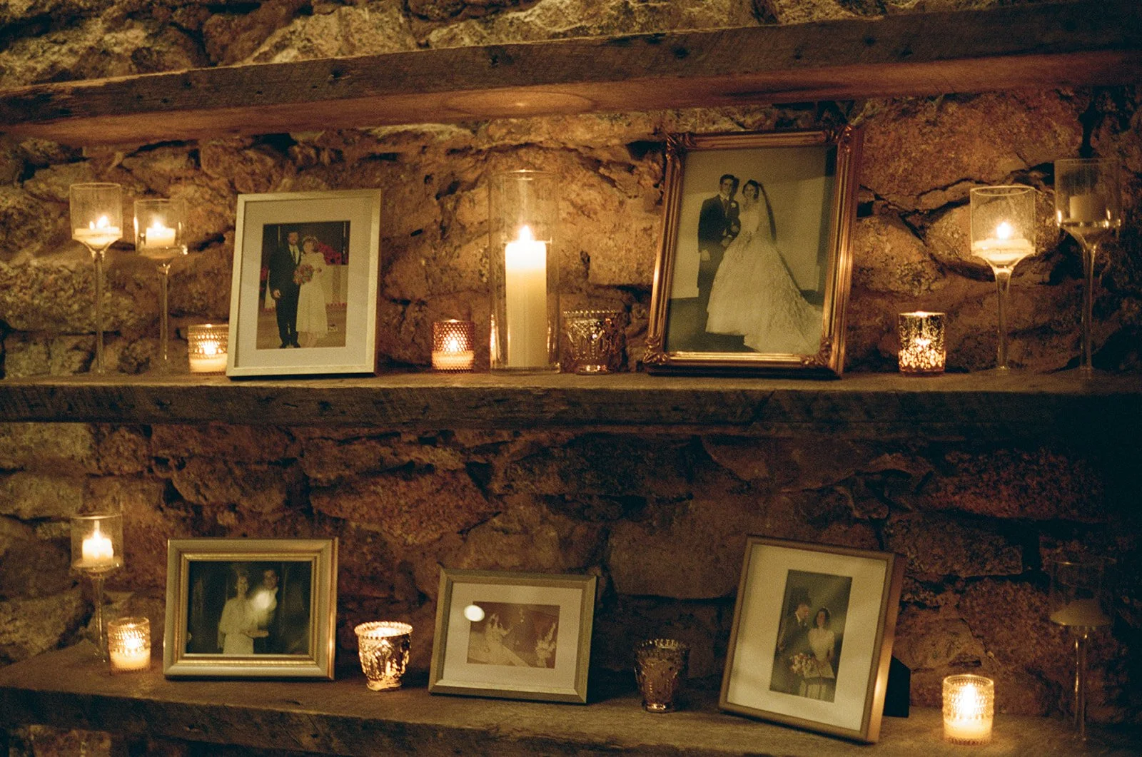 A rustic stone fireplace with two wooden shelves decorated with framed wedding photos and lit candles. potters craft cider charlottesville va