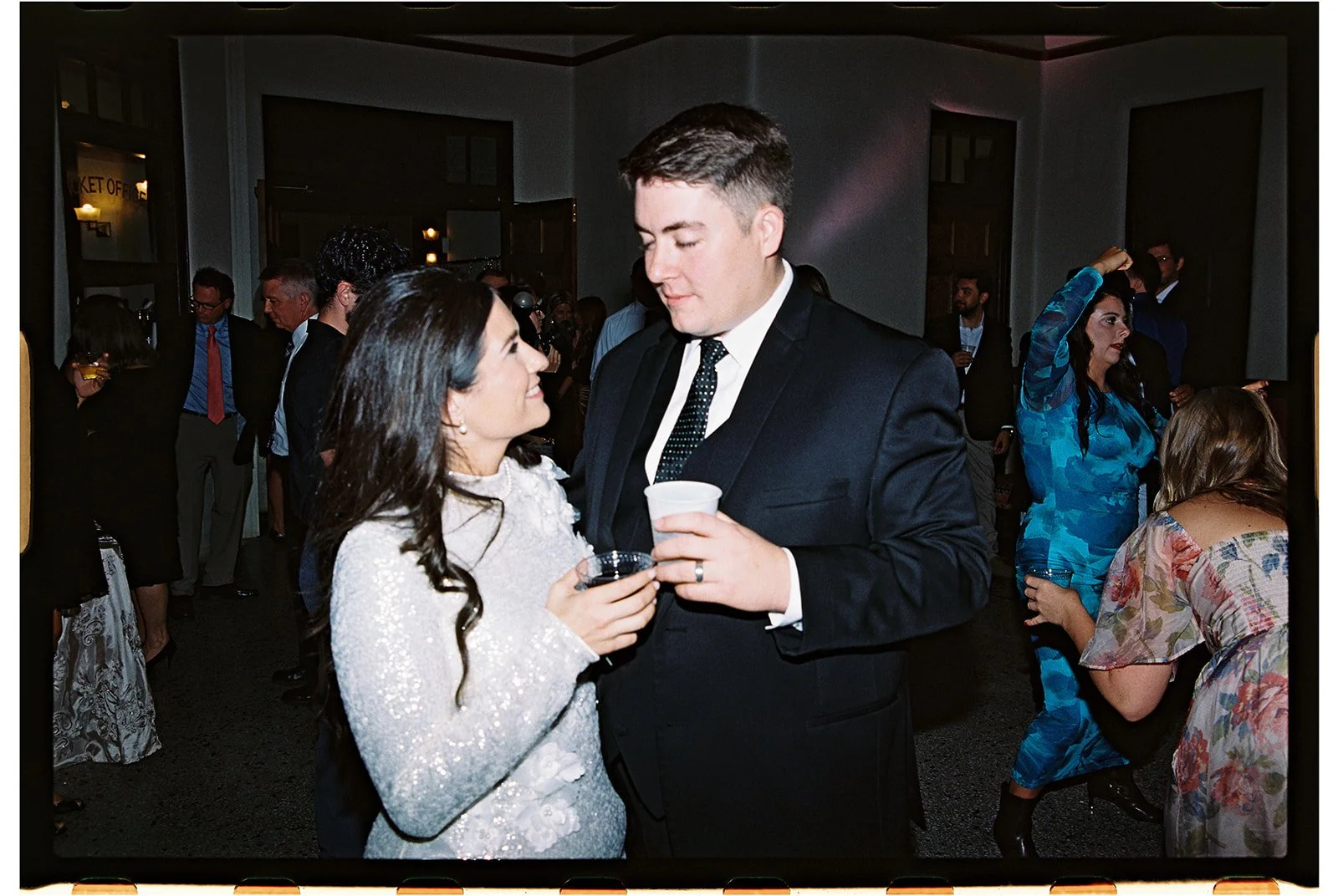 A man and woman in formal attire holding drinks and standing close together at a crowded indoor event, looking at each other. The woman wears a white sequin dress, and the man is in a dark suit with a black tie. roanoke wedding film photographer VA