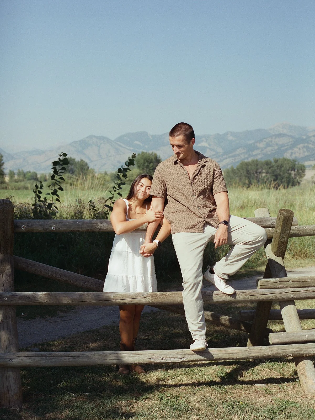 A young man and woman standing outdoors near a wooden fence, holding hands, with mountains and green fields in the background. bozeman montana engagement photographer film