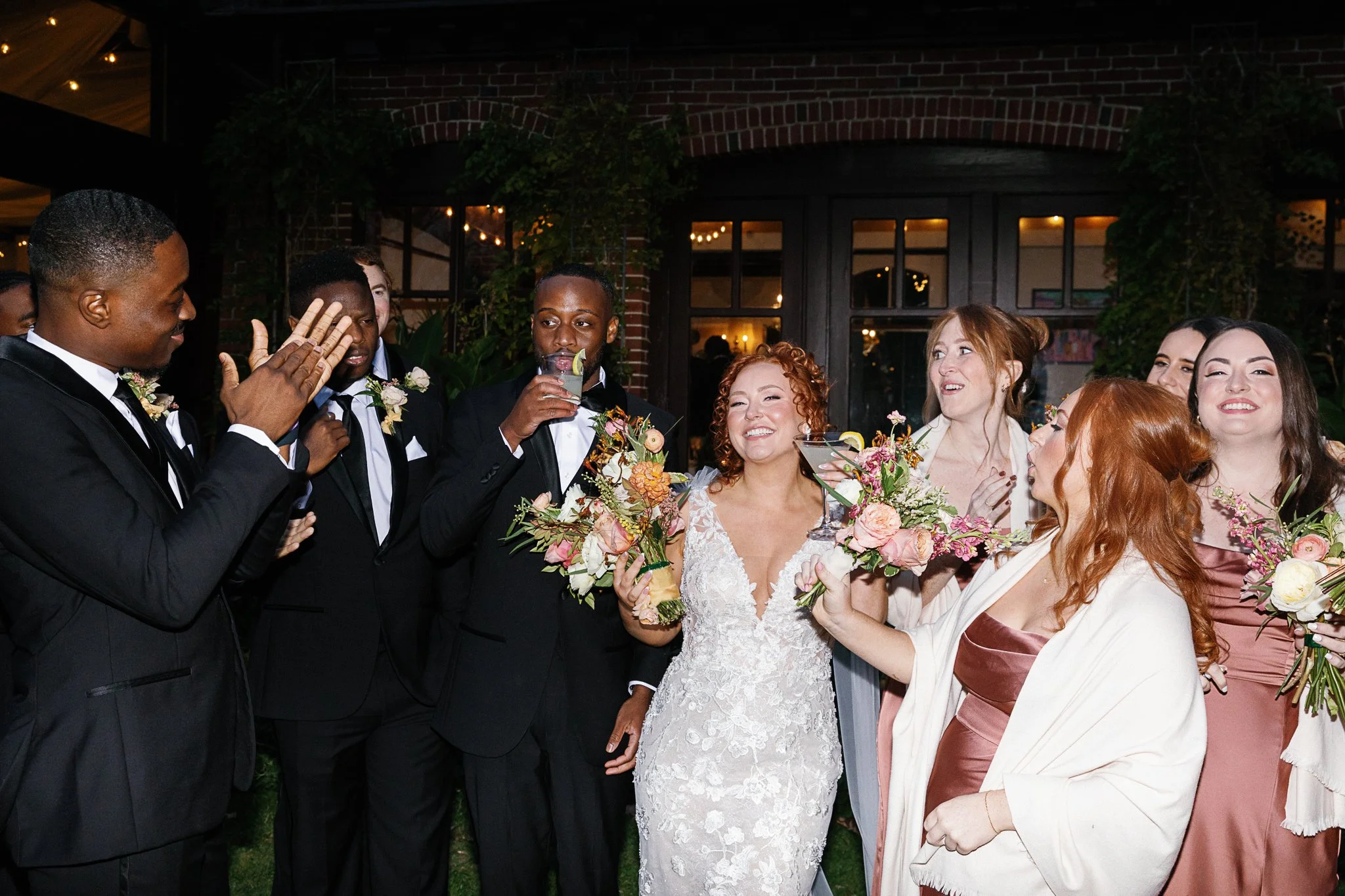 Wedding party celebrating outdoors with bride, bridesmaids holding bouquets, and groomsmen in tuxedos enjoying drinks, smiling and clapping.
