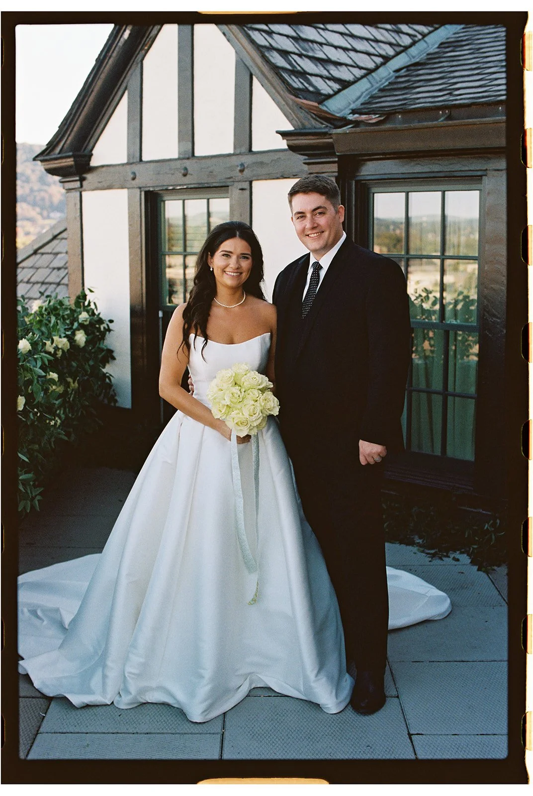 A bride and groom standing together on a balcony, smiling. The bride is wearing a white wedding dress and holding a bouquet of white roses. The groom is dressed in a black tuxedo with a tie. The background shows a house with large windows and a sloped roof, and some greenery.