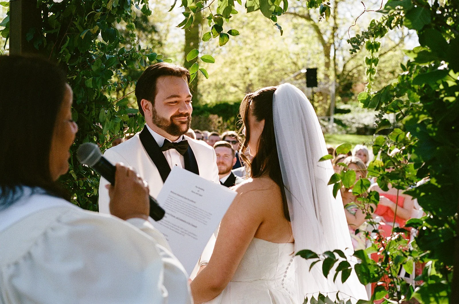 A couple getting married outdoors, exchanging vows in front of an officiant holding a microphone and notes, with guests seated on the side, surrounded by greenery. Gramercy Mansion Wedding Film Photographer Maryland Baltimore