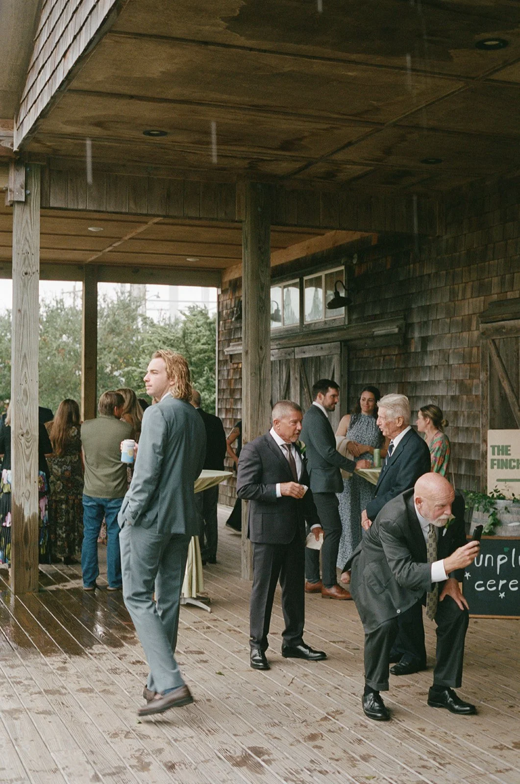 People attending a wedding reception on a wooden deck, some standing and chatting, wearing formal and semi-formal attire, with a rustic barn in the background. ocracoke outerbanks wedding film photographer
