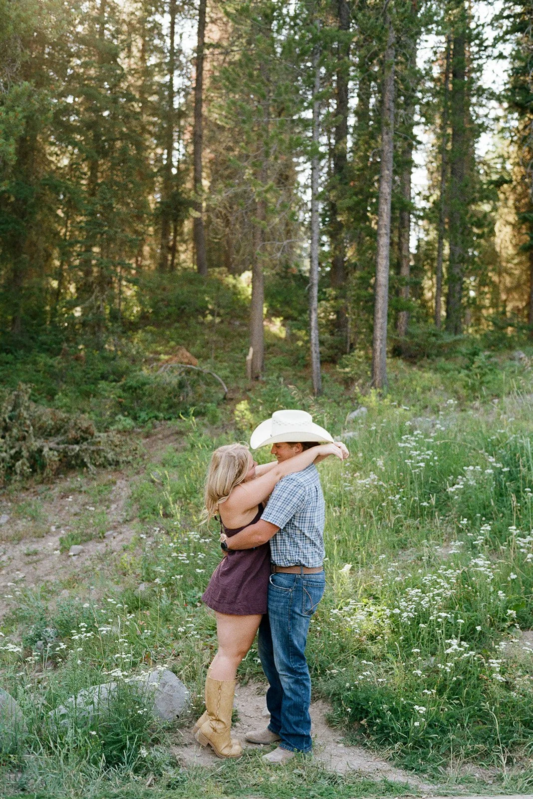 A couple hugging in a forest clearing, woman wearing tan cowboy boots and a purple dress, man wearing a cowboy hat and plaid shirt, surrounded by tall trees and wildflowers. hyalite reservoir bozeman montana engagement shoot