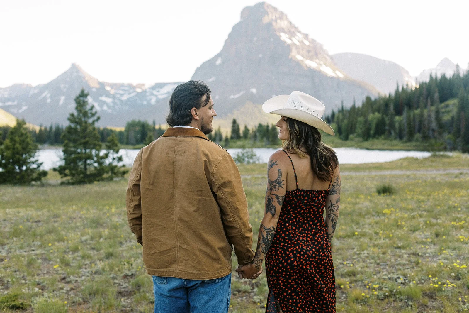 A couple holding hands, standing in a natural landscape with a lake and mountains in the background.