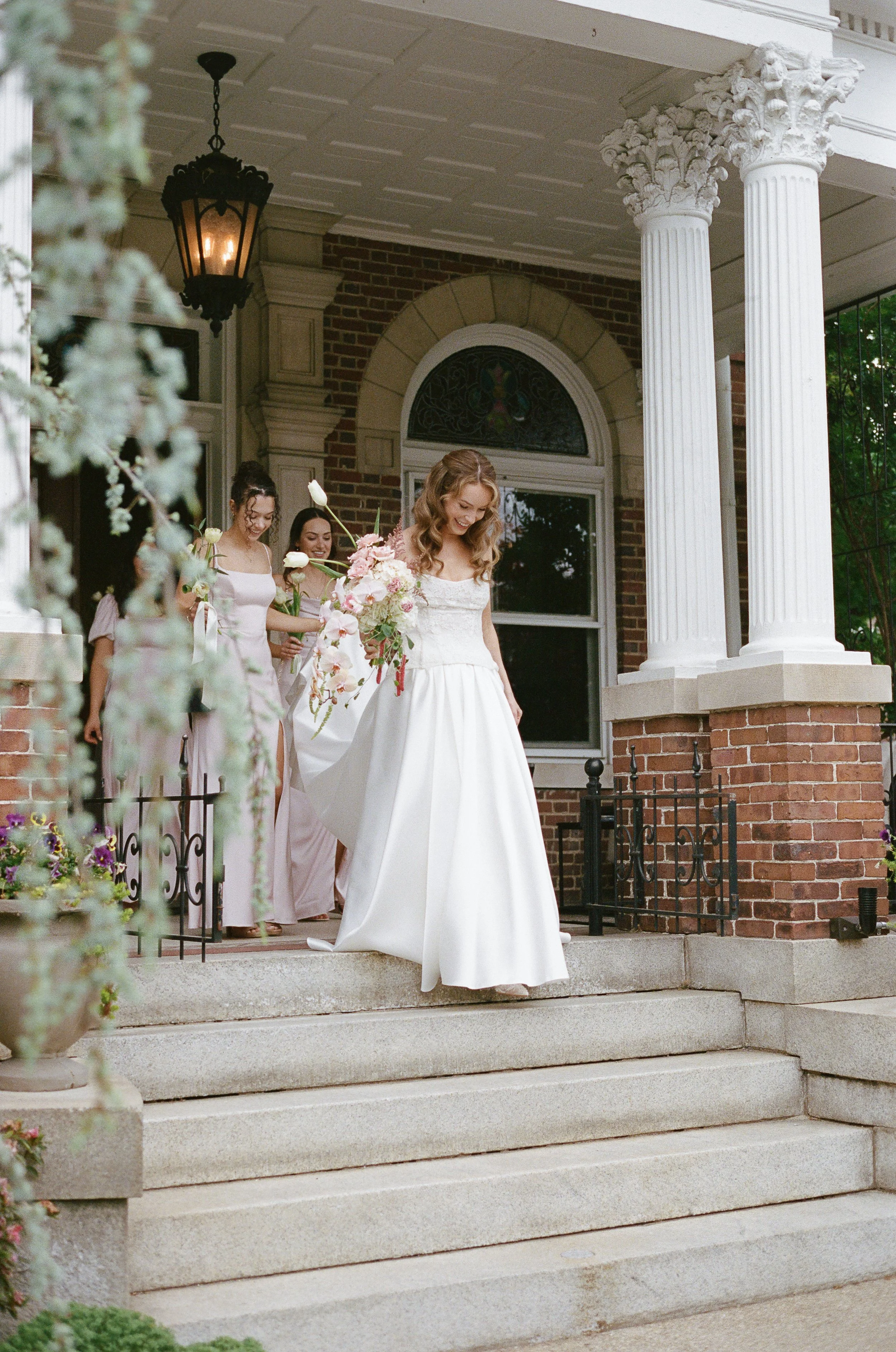 A bride in a white wedding gown holding a bouquet of pink and white flowers, standing on the steps of a brick house with white columns, with three women in pastel pink dresses behind her, one of whom is smiling.