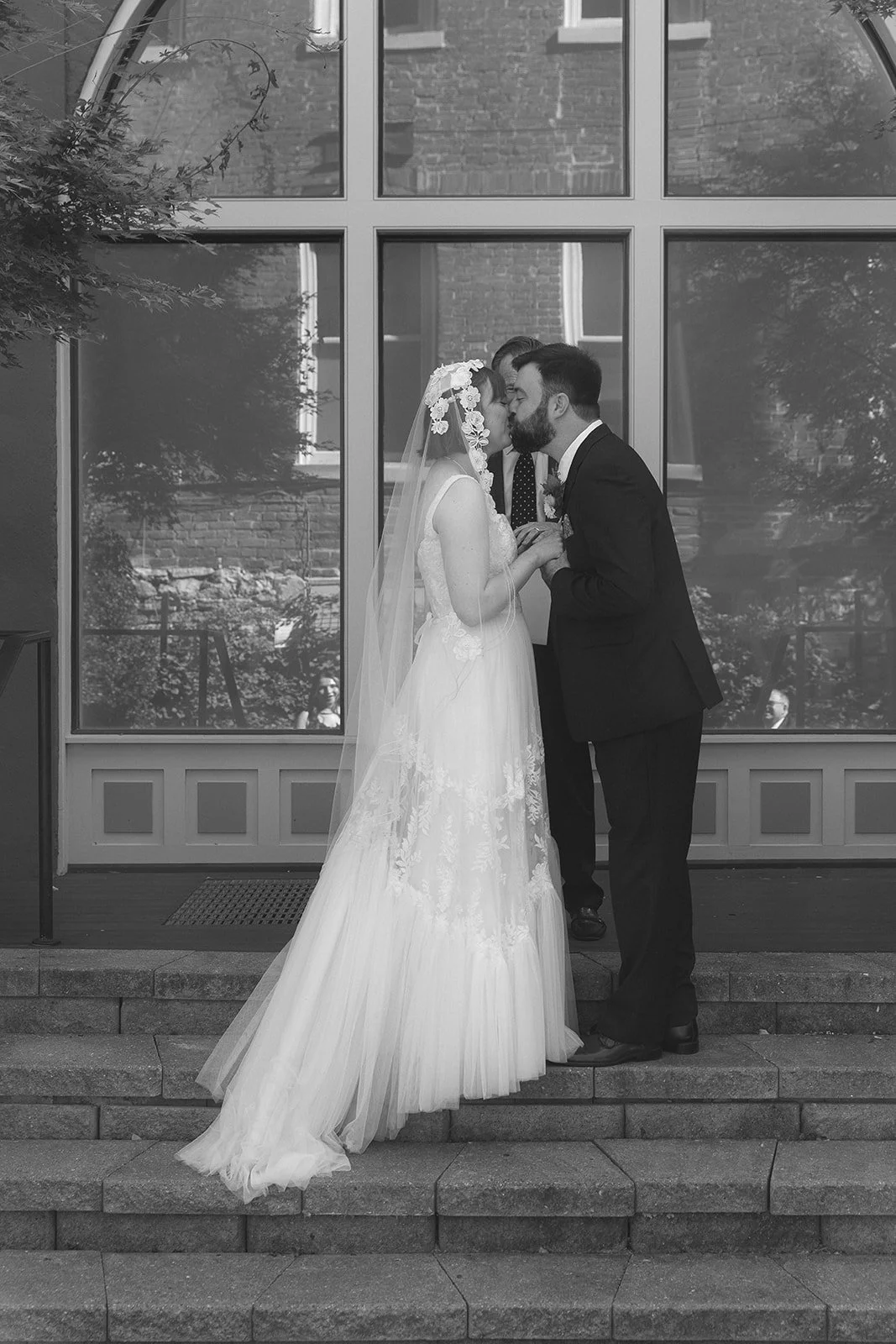A bride and groom share a kiss during their wedding ceremony on outdoor steps, with a large window and building in the background. Staunton Virginia Wedding Film Photographer.