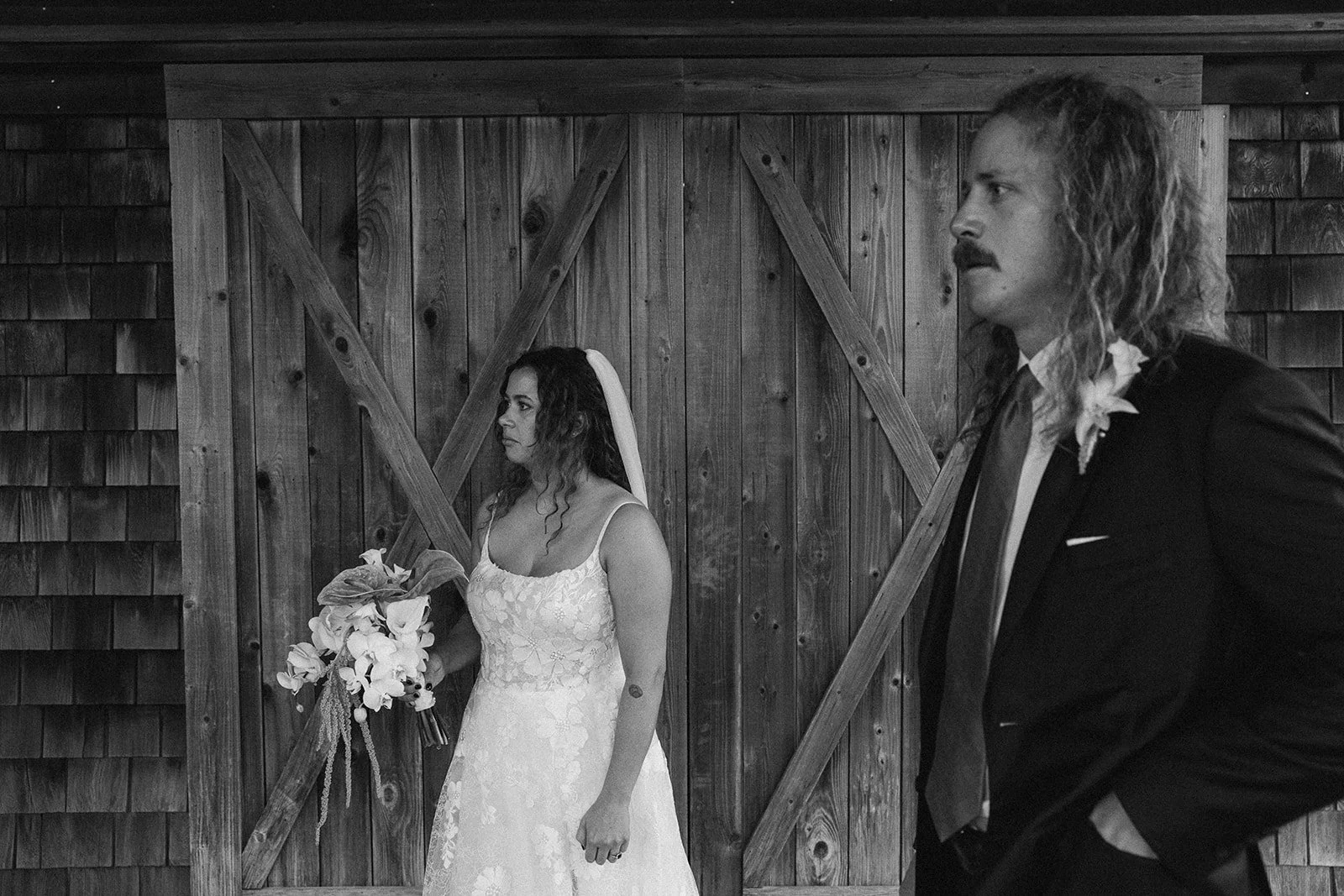 Black and white photo of a bride and groom standing apart in front of a wooden barn door, both with serious expressions. ocracoke outerbanks wedding film photographer