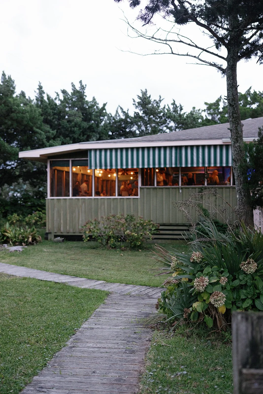 A house with a wooden exterior and a striped green and white awning, with large windows revealing an indoor gathering. The house is surrounded by green grass, plants, and trees, and there is a curved wooden pathway leading up to it during dusk or eve