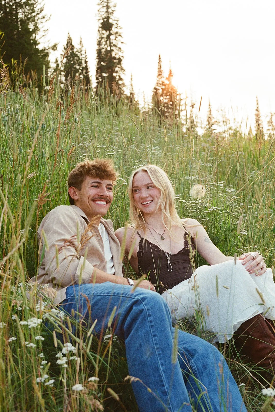 A young man and woman sitting in a grassy field during sunset, smiling and looking at each other. bozeman montana engagement photographer