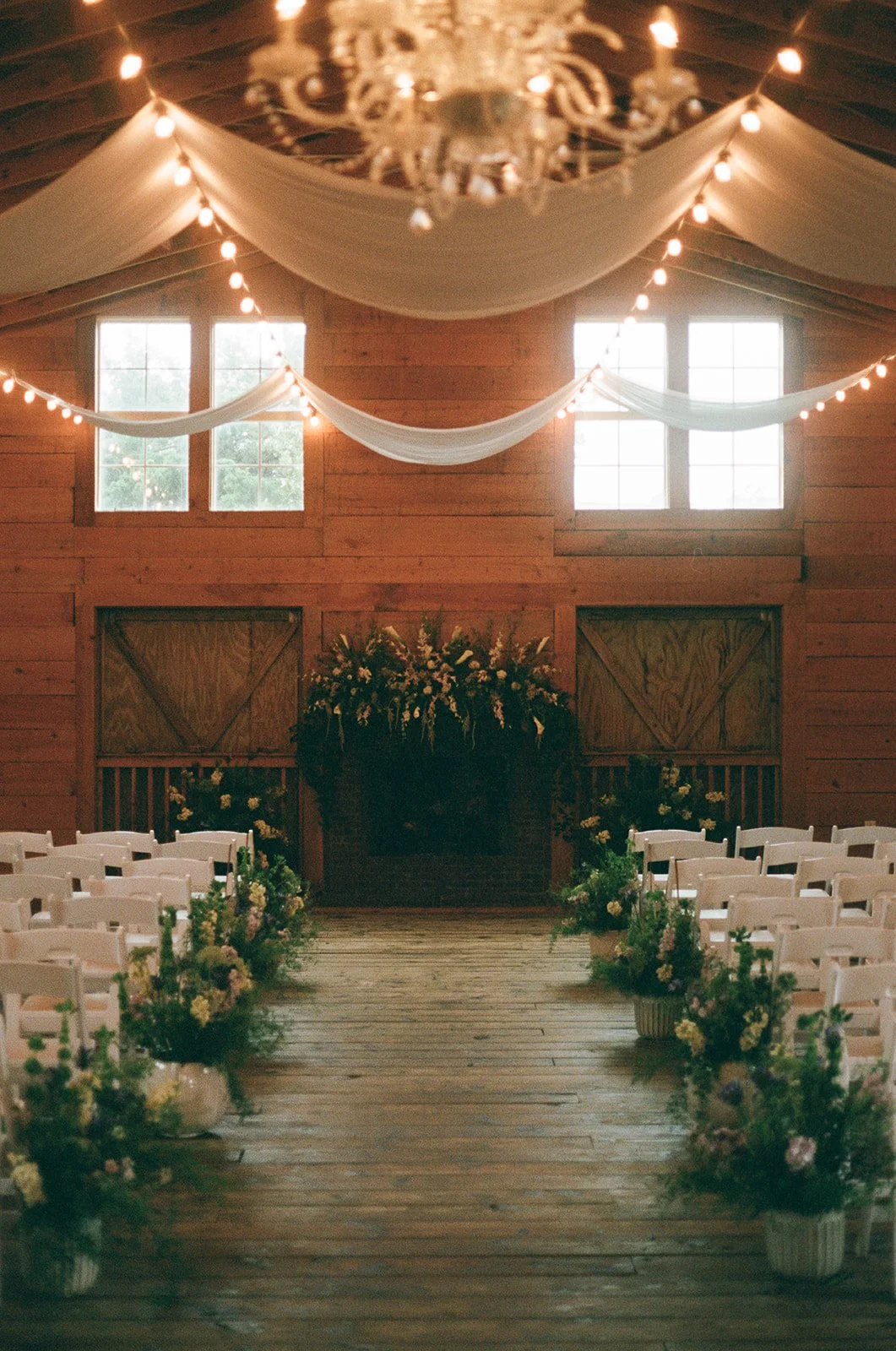  Interior of a rustic wedding ceremony setup in a wooden barn with rows of white chairs, floral arrangements along the aisle, fabric drapery, string lights, and high windows. ocracoke outerbanks wedding film photographer