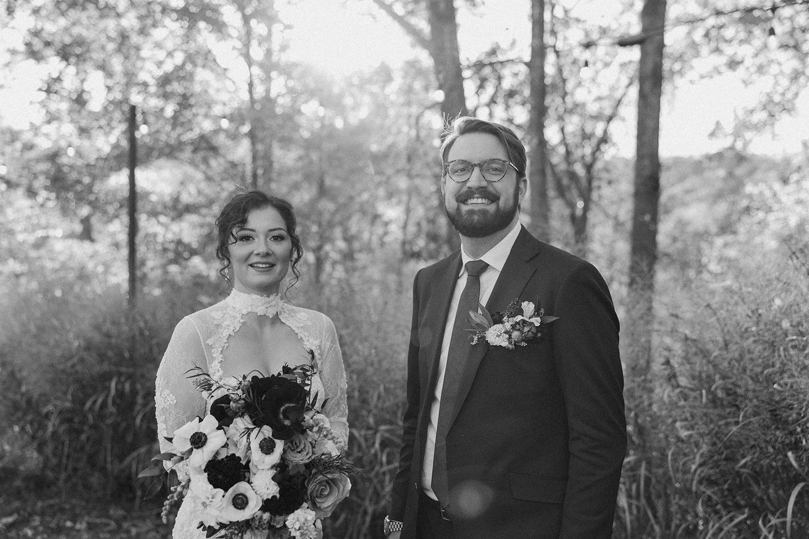 Black and white photo of a bride and groom outdoors, standing in a wooded area with trees in the background, both smiling and dressed in wedding attire. potters craft cider charlottesville va