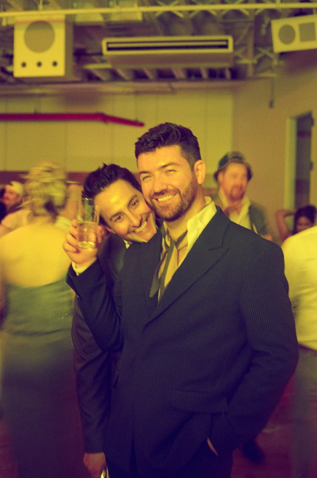 A man and woman smiling, standing close together, at a lively social event or party, with other people in the background.