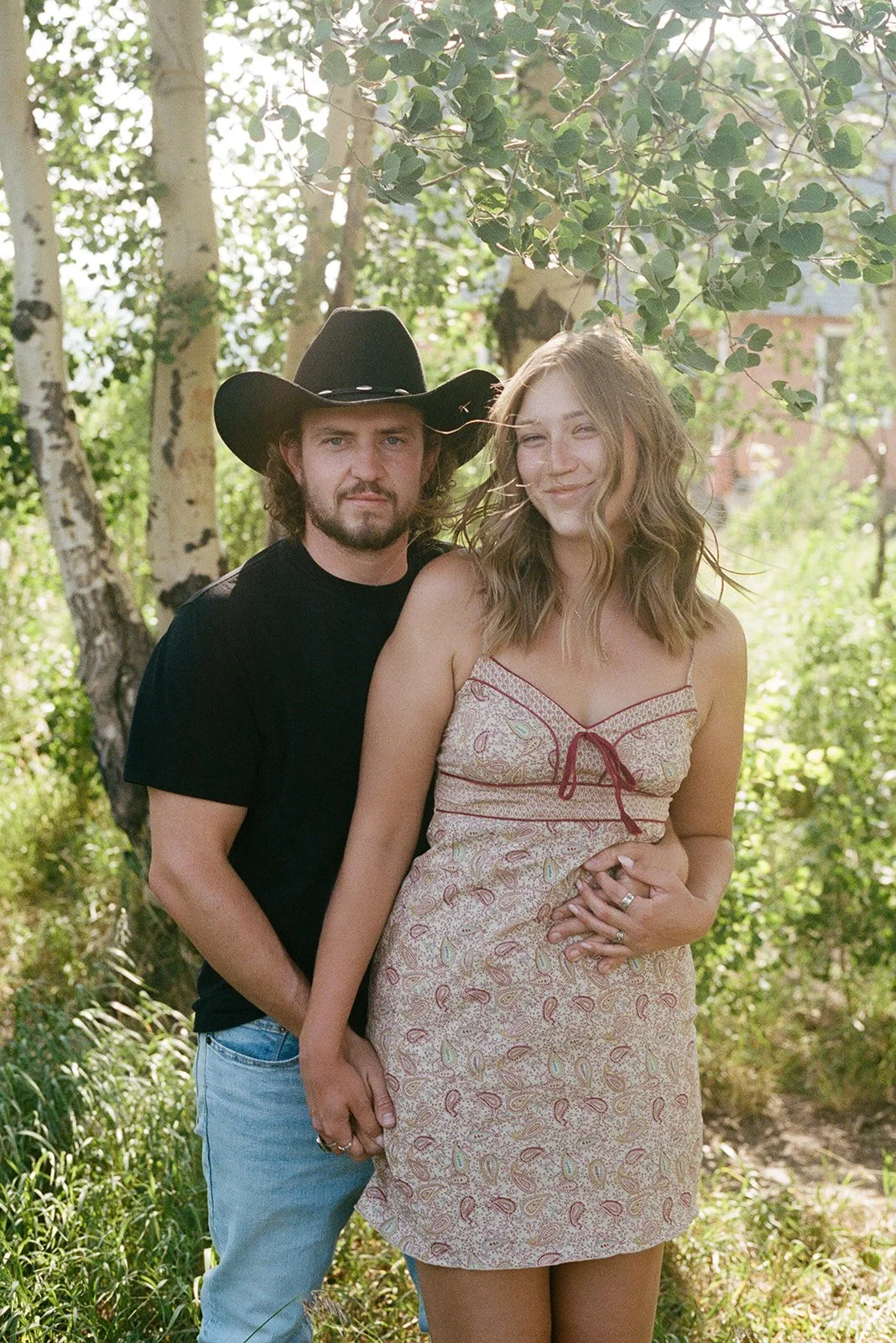 A man wearing a black cowboy hat and black t-shirt standing behind a woman in a patterned sundress in a park with green trees. mormon row jackson hole wyoming engagement shoot