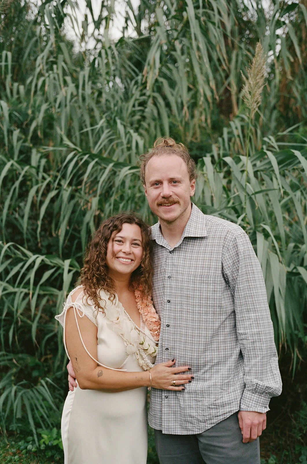 A smiling couple standing close together outdoors in front of tall green plants, with the woman wearing a light-colored dress and the man in a checkered shirt. ocracoke outerbanks wedding film photographer