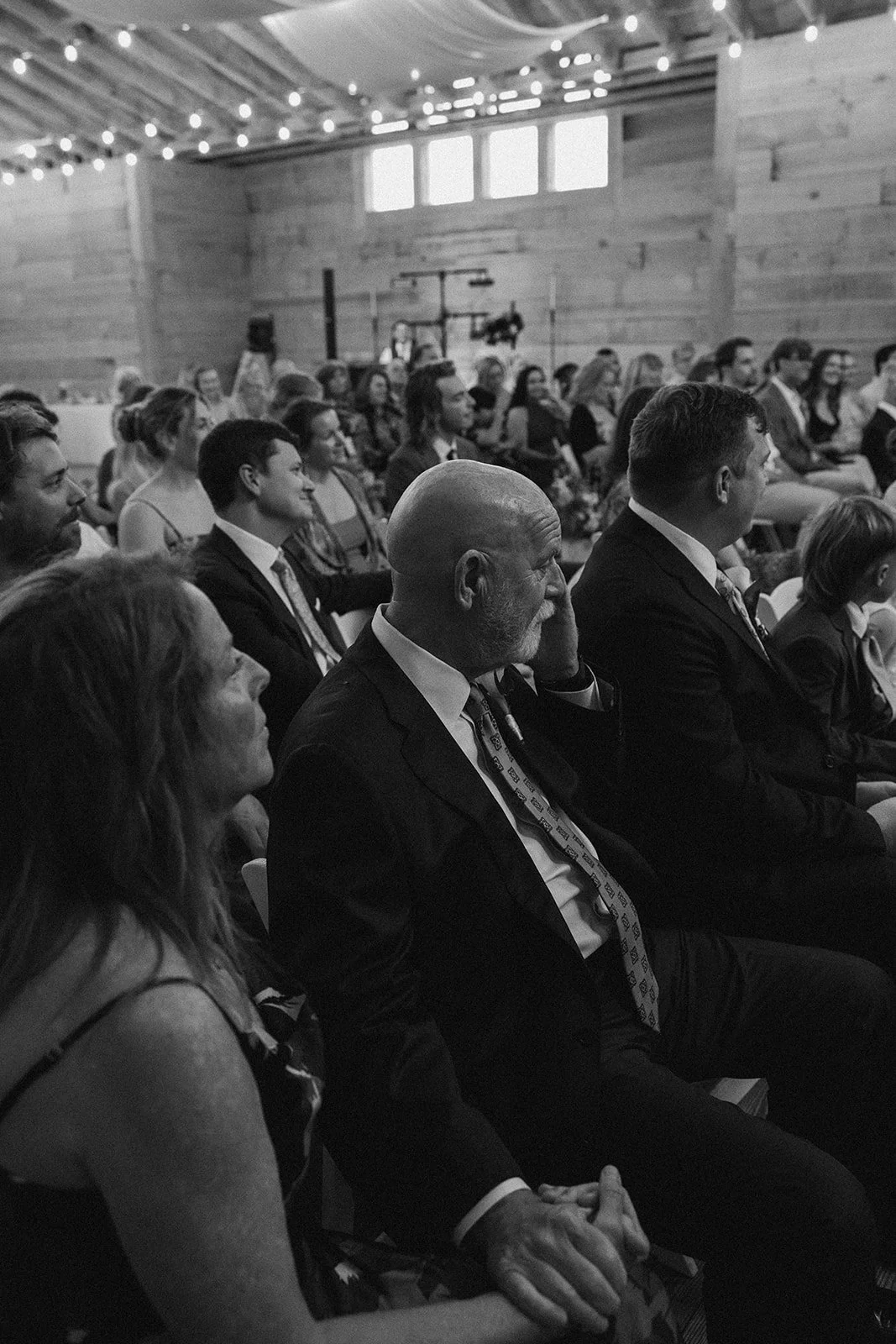 People seated in a rustic event hall watching a presentation or performance, decorated with string lights and wood walls. ocracoke outerbanks wedding film photographer