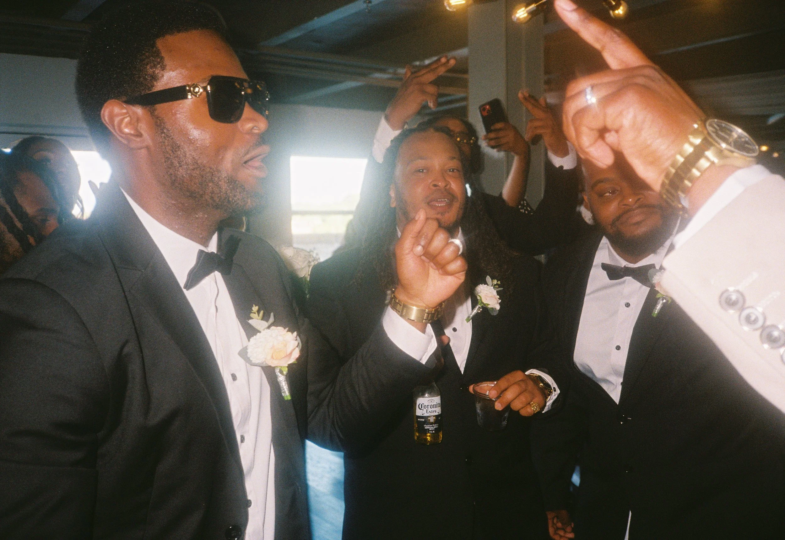 Group of men in tuxedos celebrating, some holding drinks and raising their hands, wearing boutonnieres, indoors. Roanoke Virginia