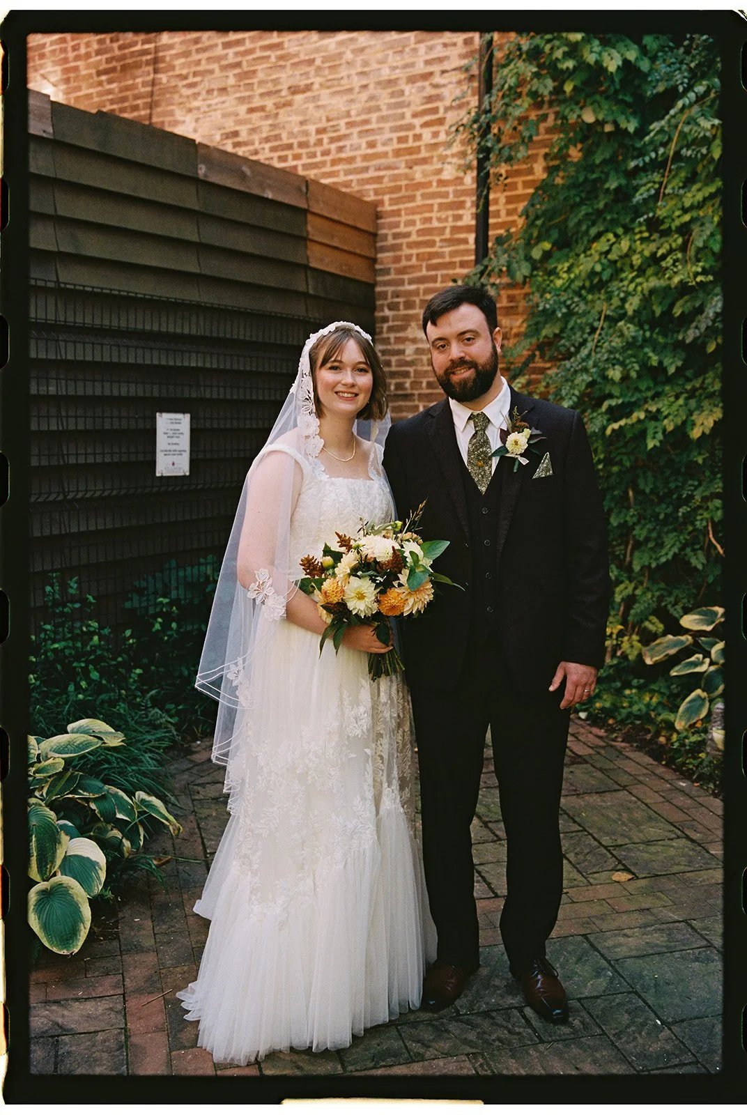 A bride and groom standing outdoors on a brick pathway with green plants and ivy in the background. The bride is holding a bouquet of flowers and wearing a white lace wedding dress with a veil. The groom is dressed in a black suit with a patterned ti