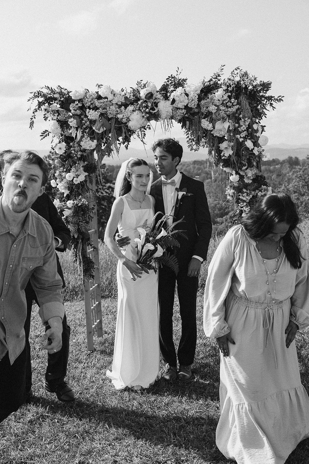 Black and white photo of a wedding ceremony outdoors with a floral arch and several people, including a bride and groom standing under the arch, with others nearby. Lexington Virginia Wedding Film Photographer