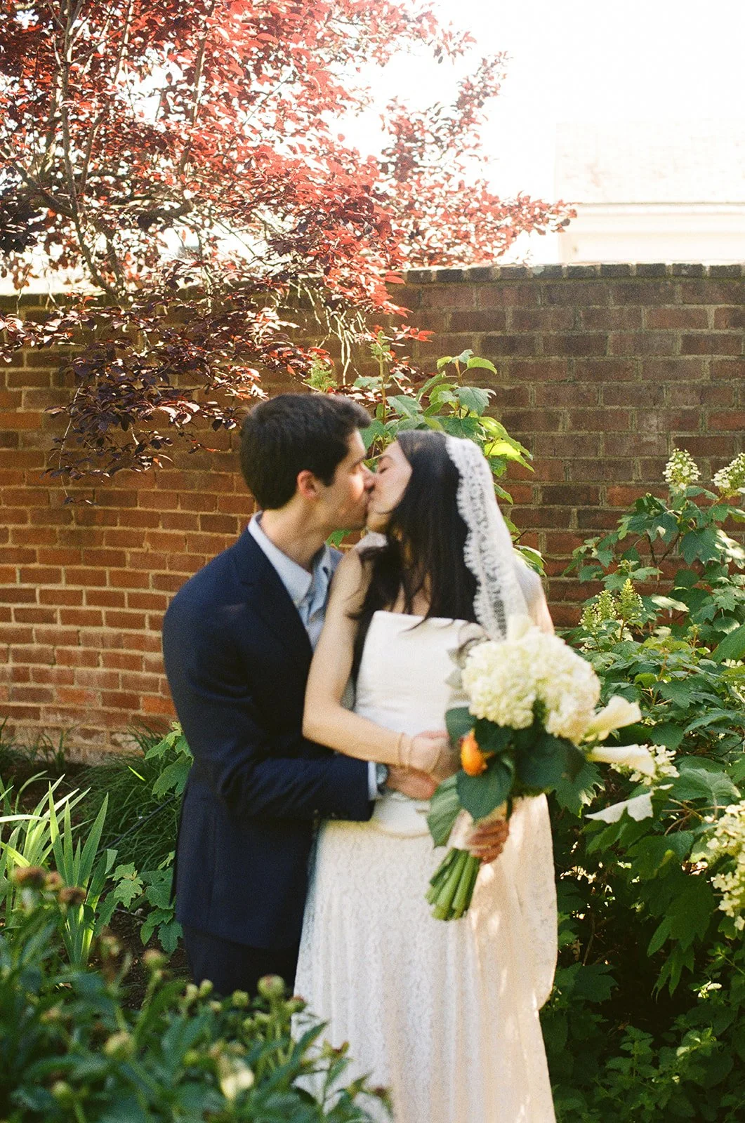 A couple in wedding attire sharing a kiss outdoors, with a brick wall and lush green plants in the background, and the woman holding a bouquet. charlottesville wedding