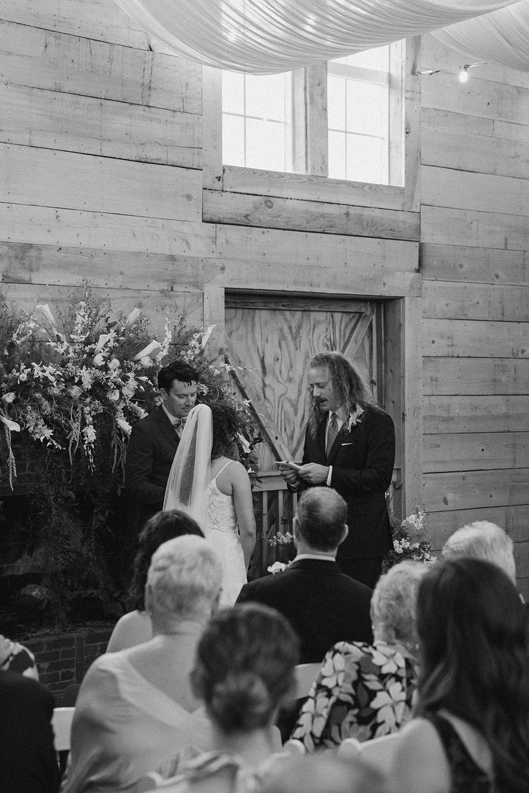 A black and white photo of a wedding ceremony with a couple standing before a officiant reading vows, inside a rustic wooden building with a floral arrangement nearby. Guests are seated and observing the ceremony. ocracoke outerbanks wedding film pho