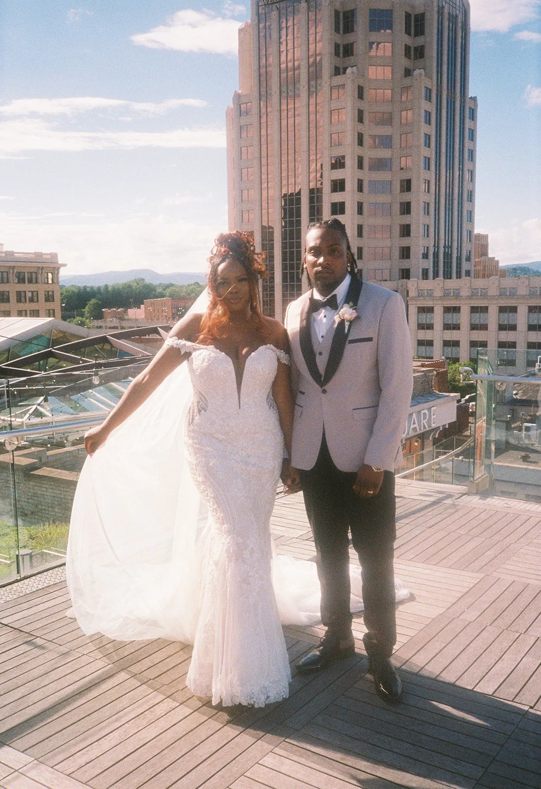 A bride and groom standing together on a rooftop terrace with city buildings in the background. The bride is wearing a white wedding dress with a veil, and the groom is in a light-colored tuxedo with a bow tie. They are holding hands. roanoke wedding
