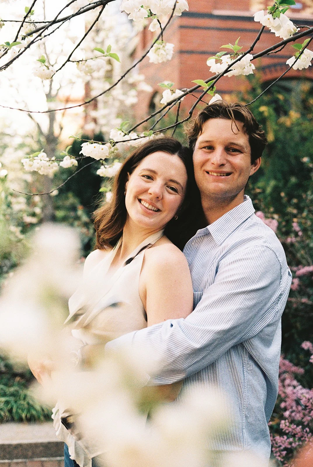 A happy couple smiling and embracing outdoors among blooming trees with white flowers, a brick building in the background.