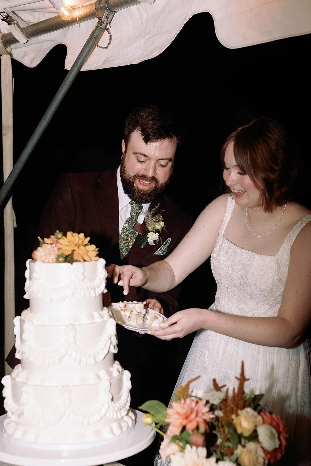 A couple cutting a wedding cake together at their wedding reception, with the bride in a white dress and the groom in a dark suit, surrounded by flowers and cake decorations. Roanoke Virginia Wedding Film Photographer.