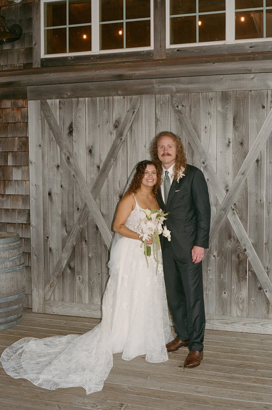 Bride and groom standing together at their wedding, in front of a rustic wooden barn door. The bride wears a white lace wedding gown and holds a bouquet, while the groom is dressed in a black suit with a white shirt and tie. outerbanks wedding film