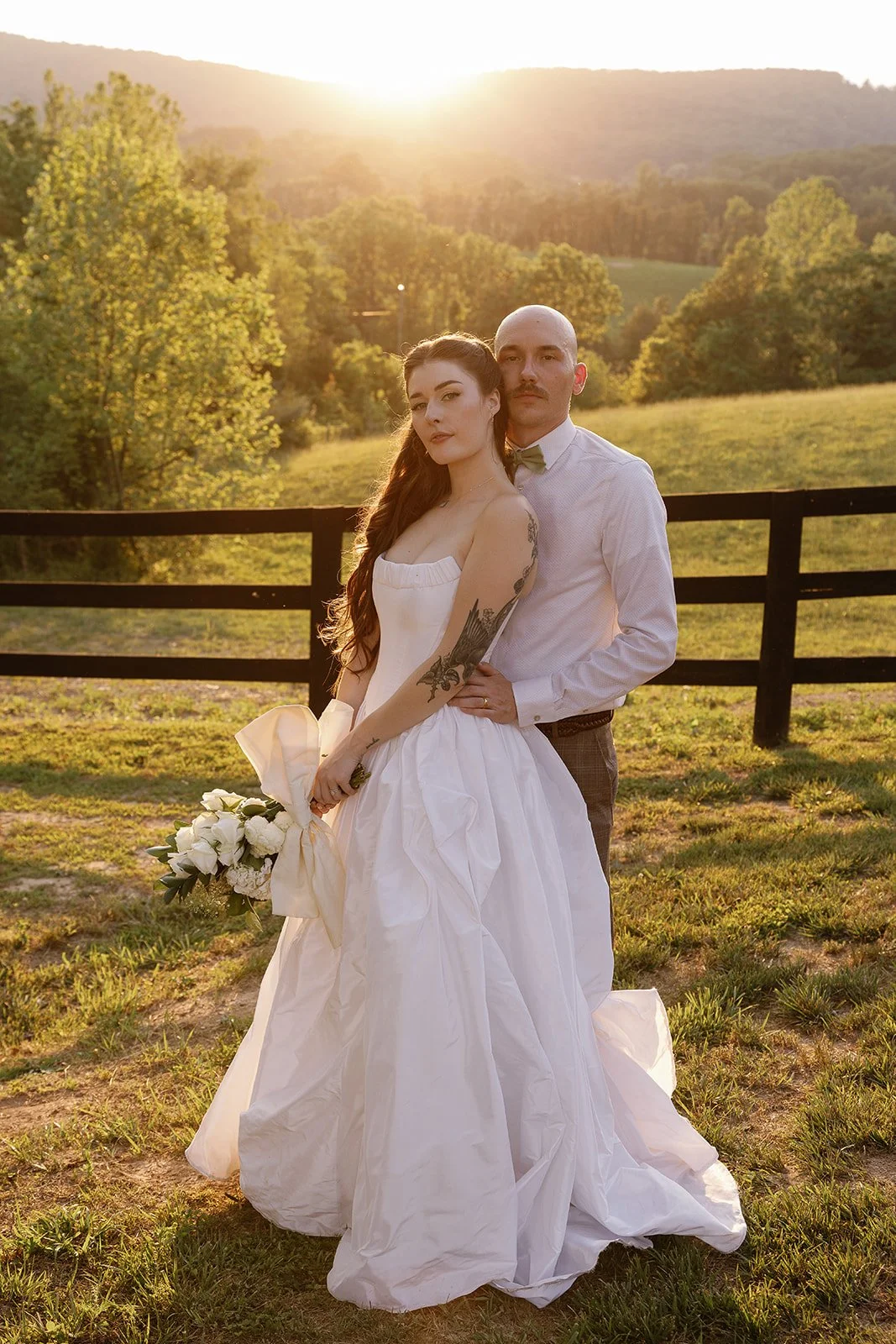 A newlywed couple poses outdoors during sunset. The bride is holding a bouquet of white flowers and wears a strapless white gown with tattoos on her arm. The groom is dressed in a white shirt and a bow tie. They stand in front of a black wooden fence with a scenic backdrop of trees, rolling hills, and the setting sun.
